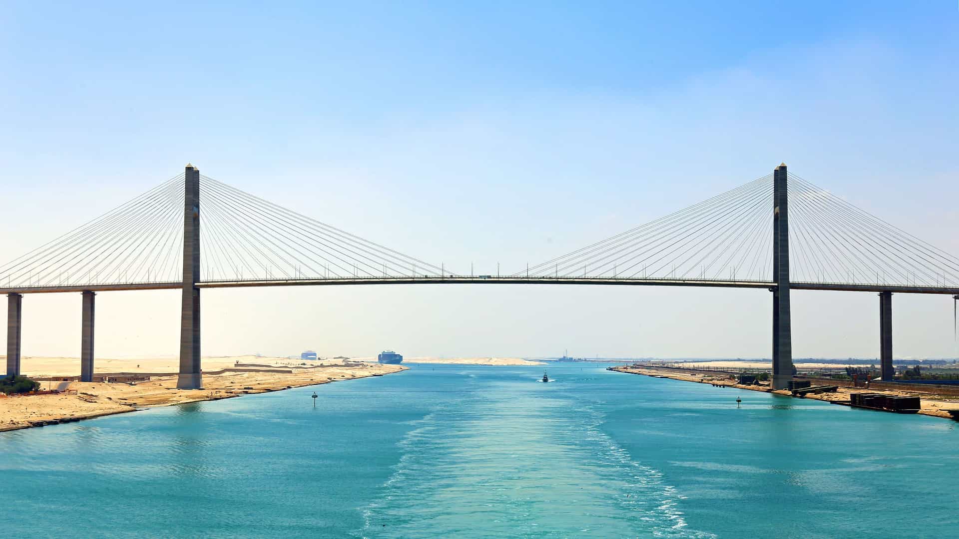 A massive suspension bridge, the El Ferdan Railway Bridge, spanning the Suez Canal in Egypt, with a large container ship visible in the distance.