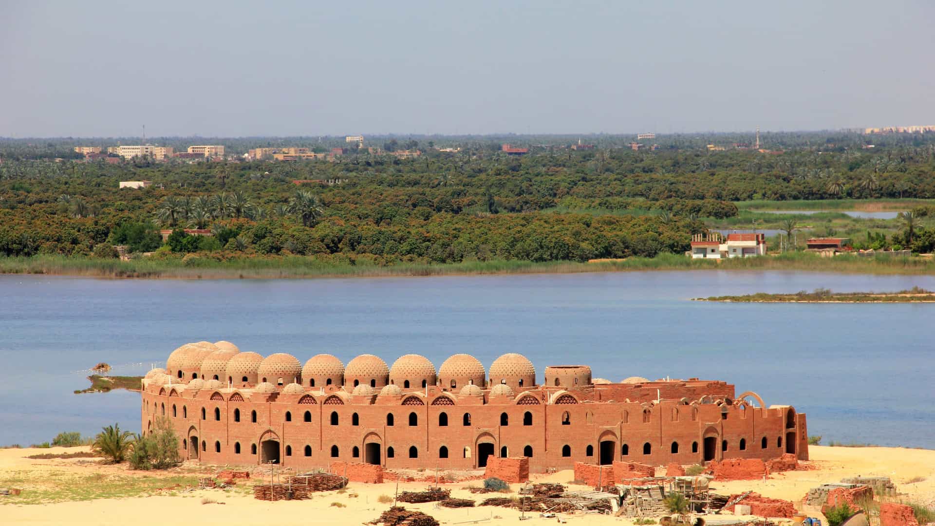 A panoramic shot of an unfinished, dome-shaped brick building on a sandy bank in Suez, Egypt, with a large body of water and a lush green forest in the background.