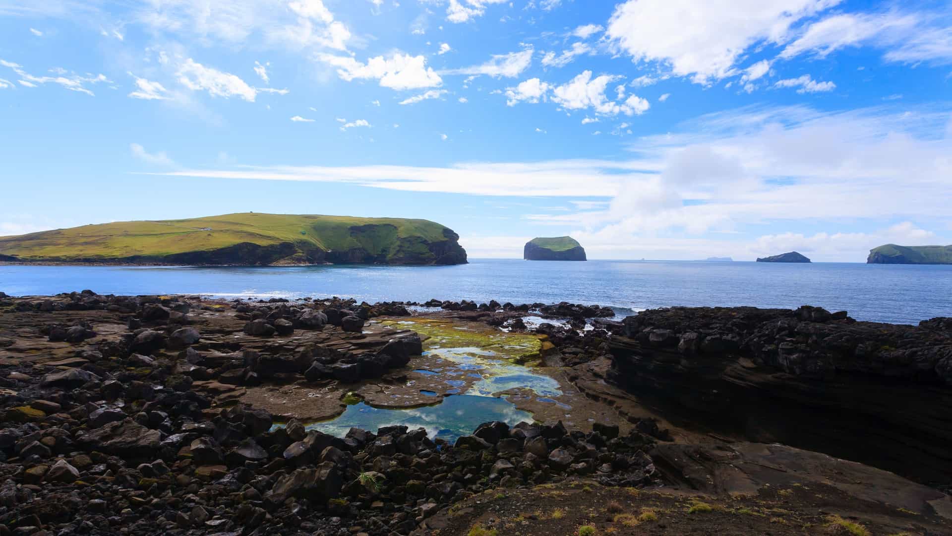 A scenic view of the volcanic Surtsey Island in Iceland, showing a rocky shoreline and calm ocean with several small, green-topped islands in the distance under a blue, cloudy sky.