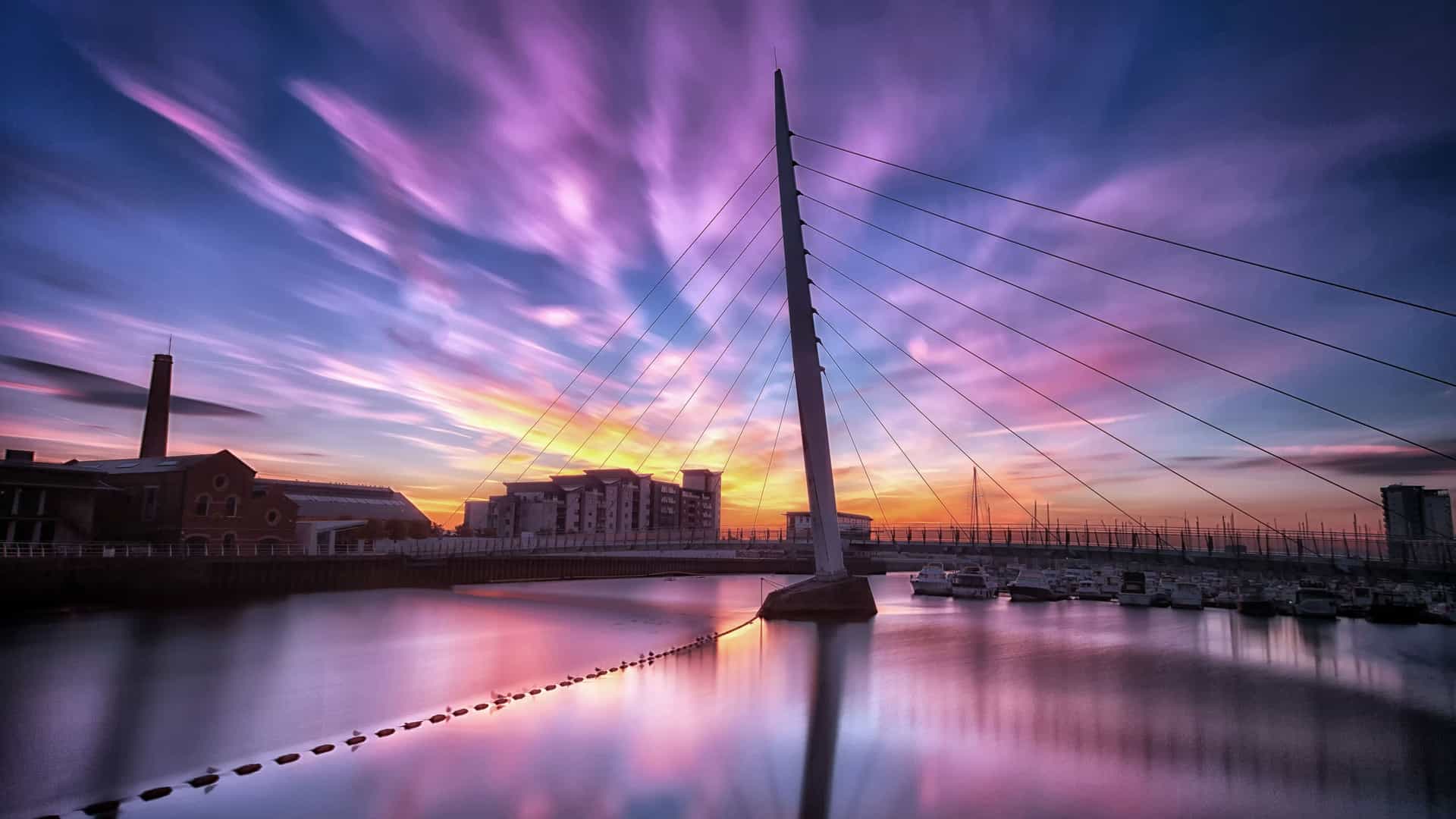 A breathtaking sunset over the Swansea Bay, Wales, with a modern suspension bridge crossing the water and vibrant colors reflecting in the marina.