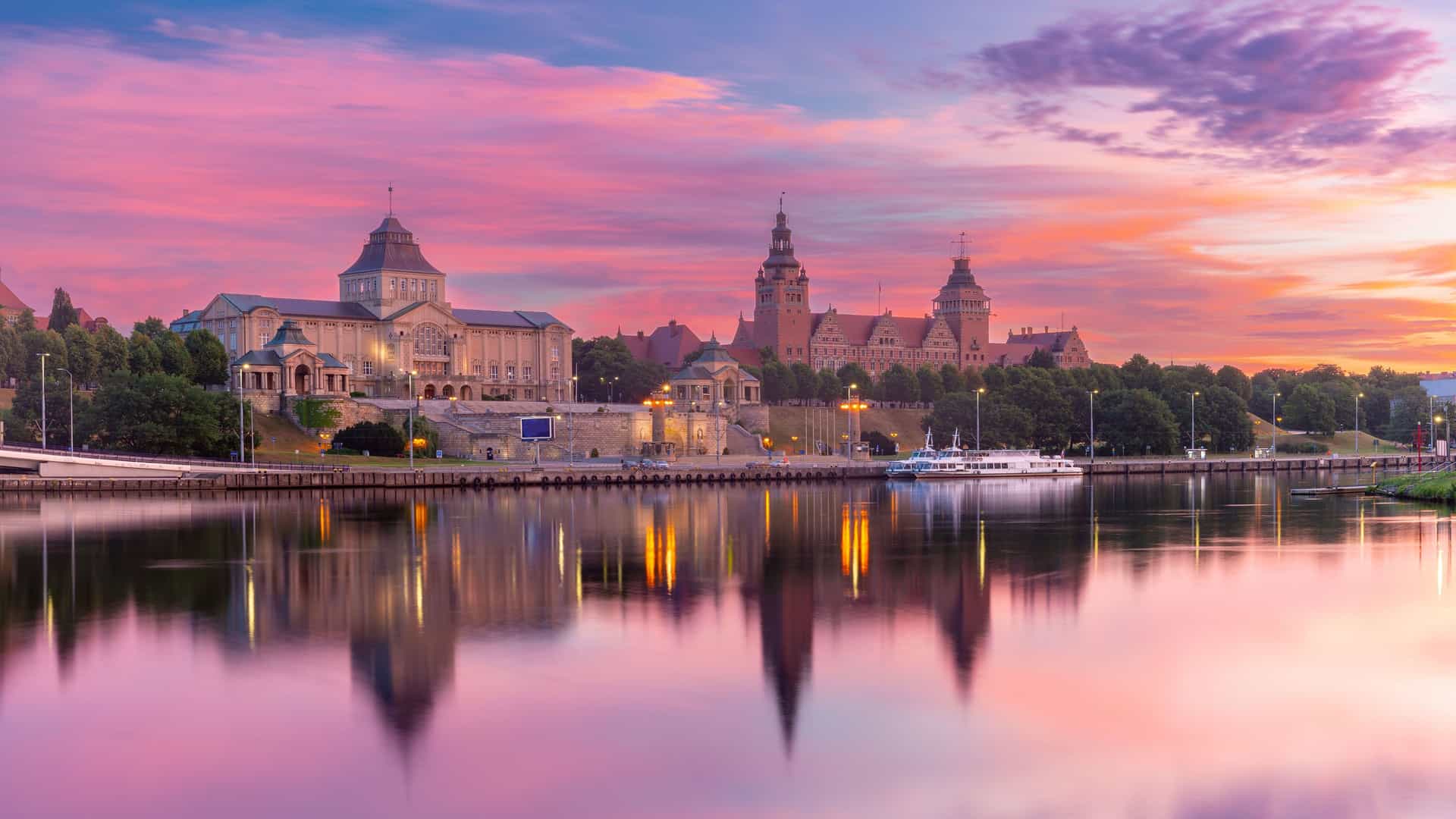 A panoramic sunset view of the waterfront in Szczecin, Poland, with the historic Chrobry Embankment and its magnificent buildings and towers reflecting beautifully in the calm Oder River. The sky is a stunning mix of pink, orange, and purple.