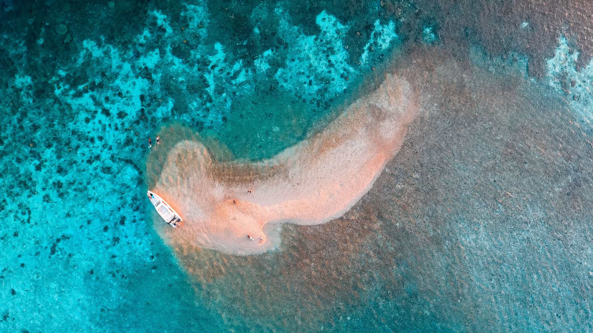 An aerial view of the Tagat Sagel Reef in Indonesia, with a small sandbar in the middle of a crystal clear turquoise sea and a small white boat and people on the sand.