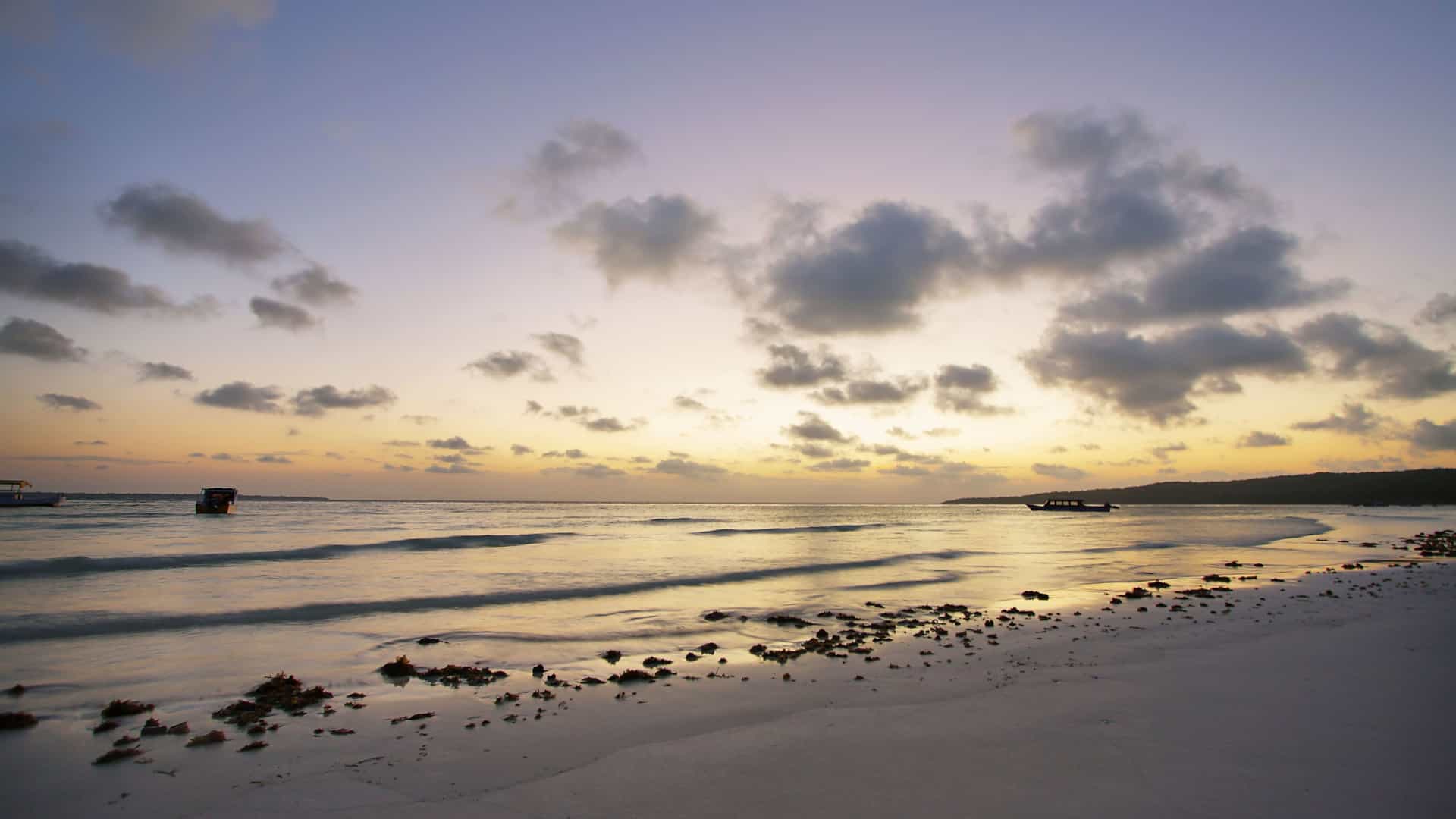 A peaceful sunset on the tropical beach of Tanjung Priok in Indonesia, with gentle waves lapping the shore under a cloudy, pastel-colored sky.