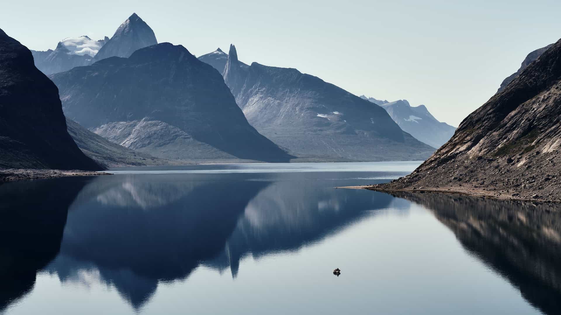 A tranquil and serene view of the Tasermiut Fjord in Greenland, with a lone boat on the calm water, which perfectly reflects the towering, dark, and rugged mountain peaks that surround the fjord.