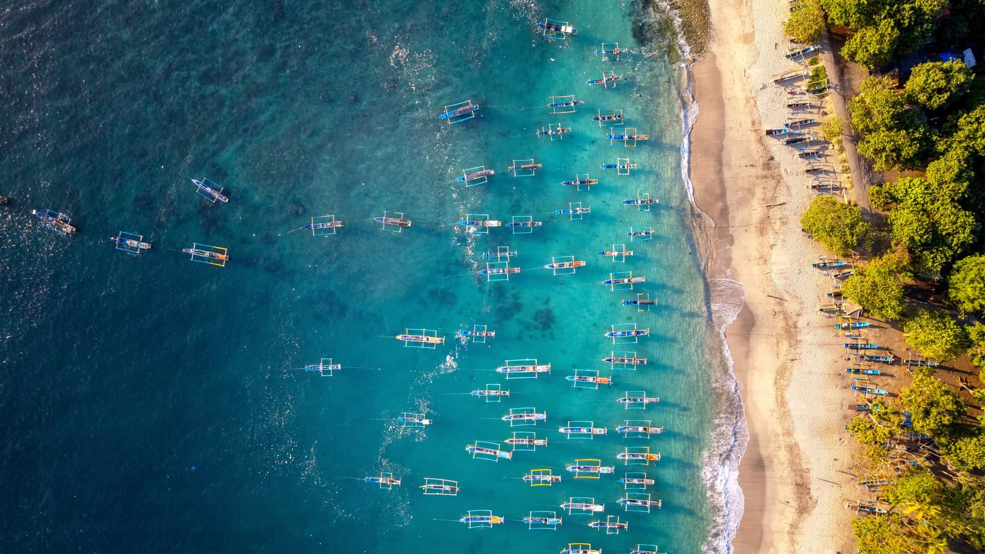 A stunning top-down aerial view of dozens of traditional wooden outrigger boats anchored in crystal-clear turquoise water along a pristine white sand beach in Tatawa Besar, Indonesia.