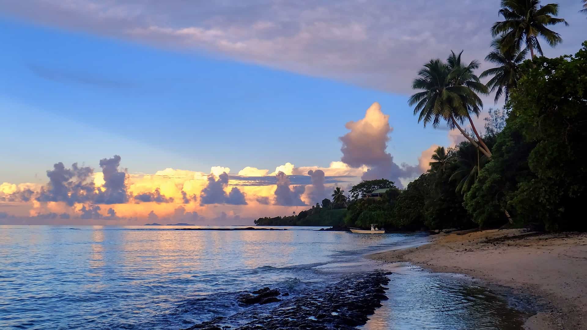 A panoramic landscape of Taveuni Island in Fiji, with a lush, green tropical jungle and palm trees sloping down to a beautiful blue coastline and distant islands under a bright, cloudy sky.