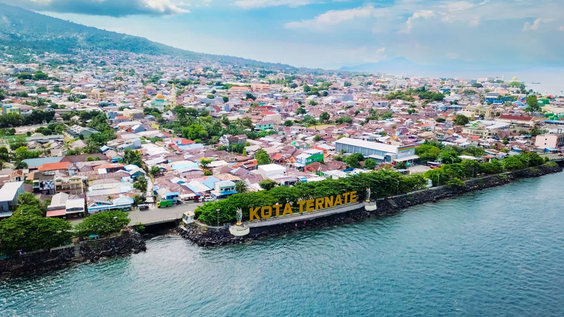 An aerial view of the coastal city of Ternate, Indonesia, with colorful houses, a waterfront, and a large "KOTA TERNATE" sign near the sea.