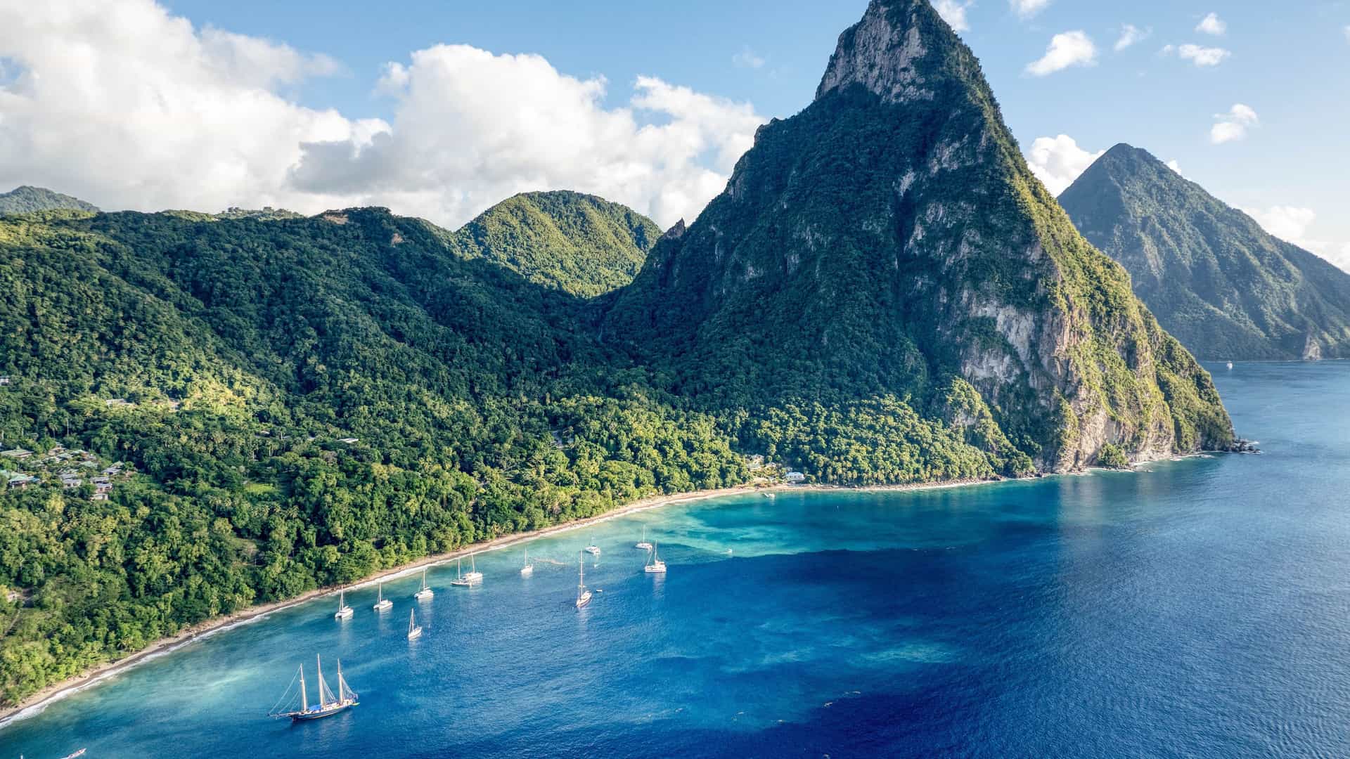 A breathtaking aerial view of the iconic Pitons, two majestic volcanic peaks, rising from the turquoise waters of the Caribbean Sea on the island of St. Lucia, with a small bay and sailboats.