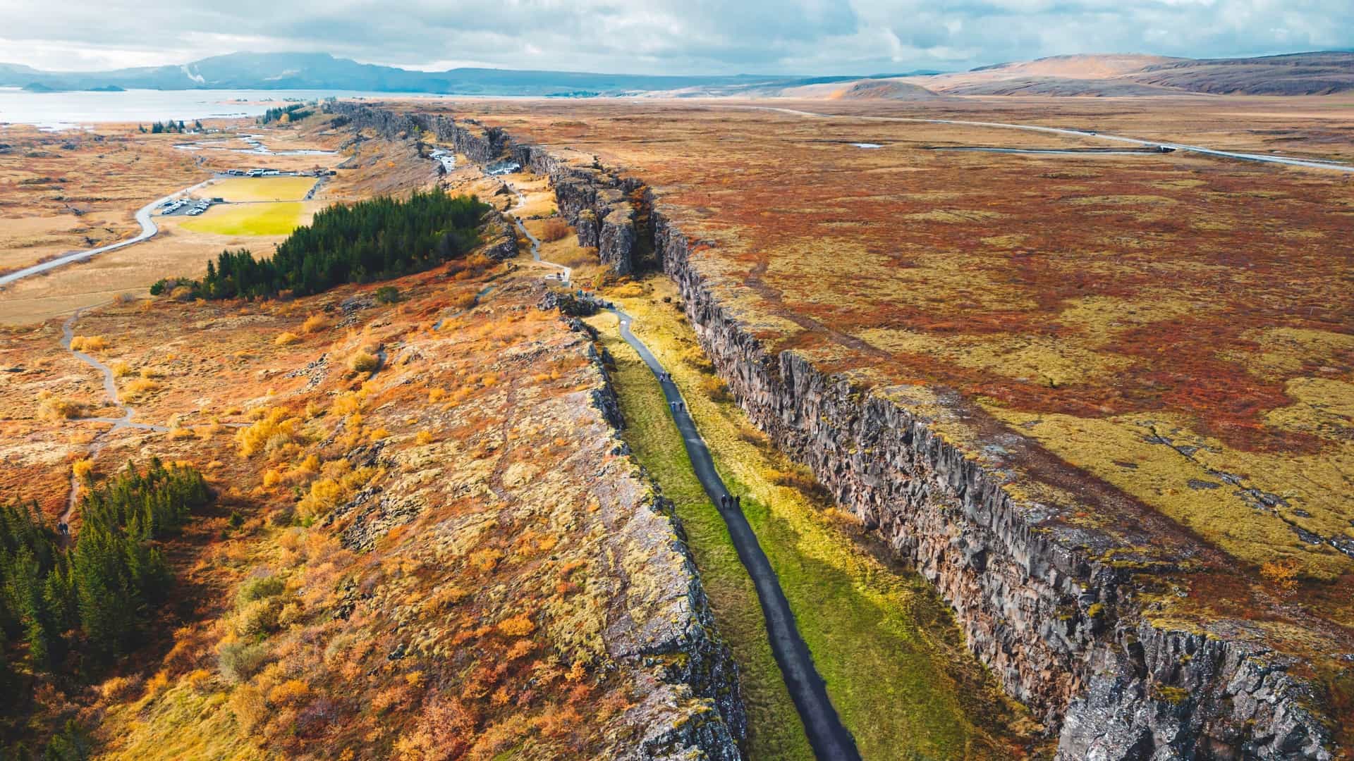 A dramatic aerial view of Þingvellir (Thingvellir) National Park in Iceland, a UNESCO World Heritage site and the location of the Mid-Atlantic Ridge. The image clearly shows the immense rift valley that separates the North American and Eurasian tectonic plates, characterized by steep cliffs, fissures, and the vast, serene waters of Þingvallavatn Lake in the background.