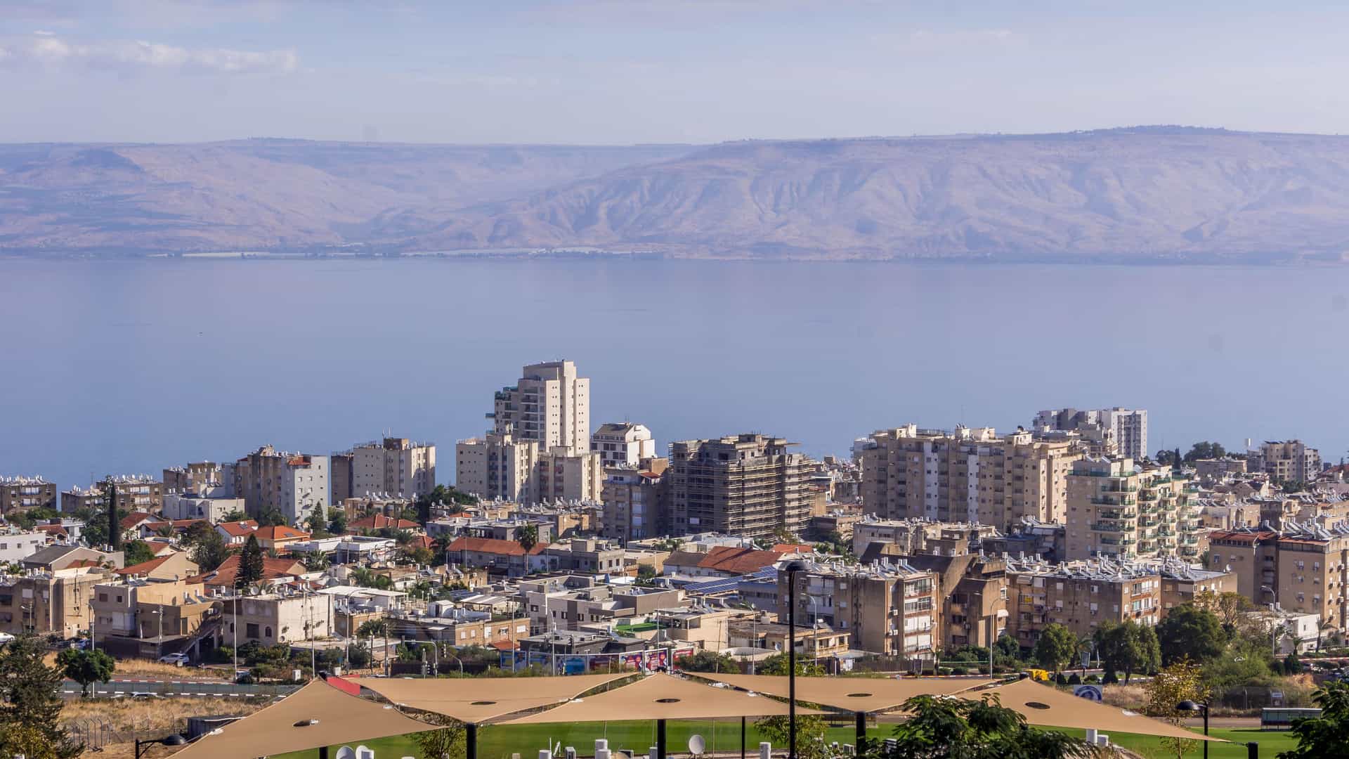"A scenic view of the city of Tiberias, Israel, showcasing a cluster of buildings along the shore of the calm Sea of Galilee, with rolling desert mountains in the distance under a blue sky.  "