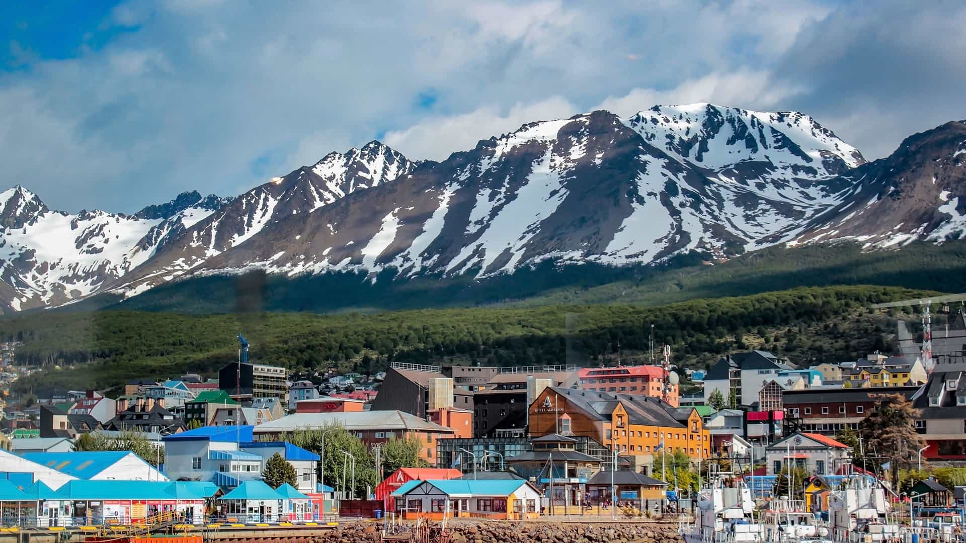 A stunning view of the colorful waterfront town of Ushuaia, Argentina, with snow-capped mountains of Tierra del Fuego National Park in the background.