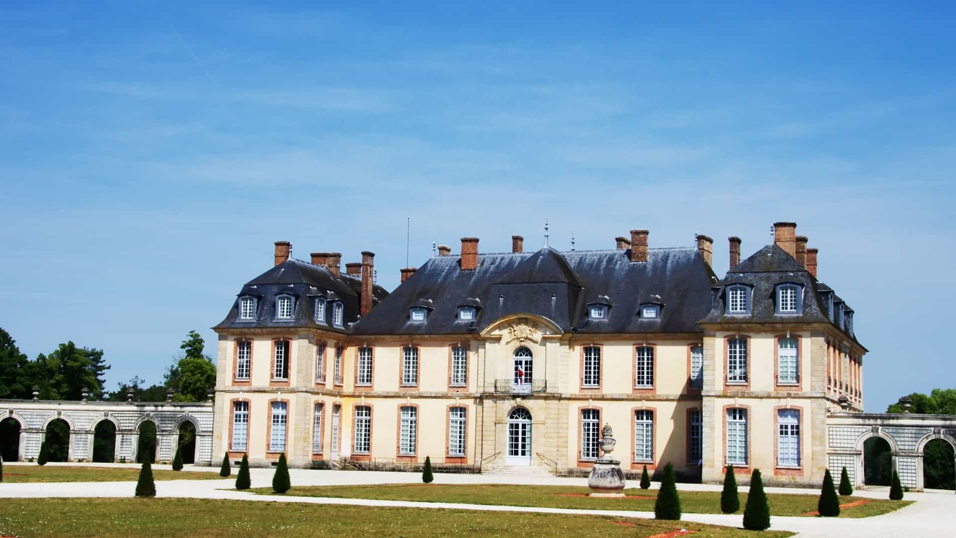 The classic, pale yellow Château de Tilly features a mansard roof and dormer windows, surrounded by a large, sunny lawn and archways.