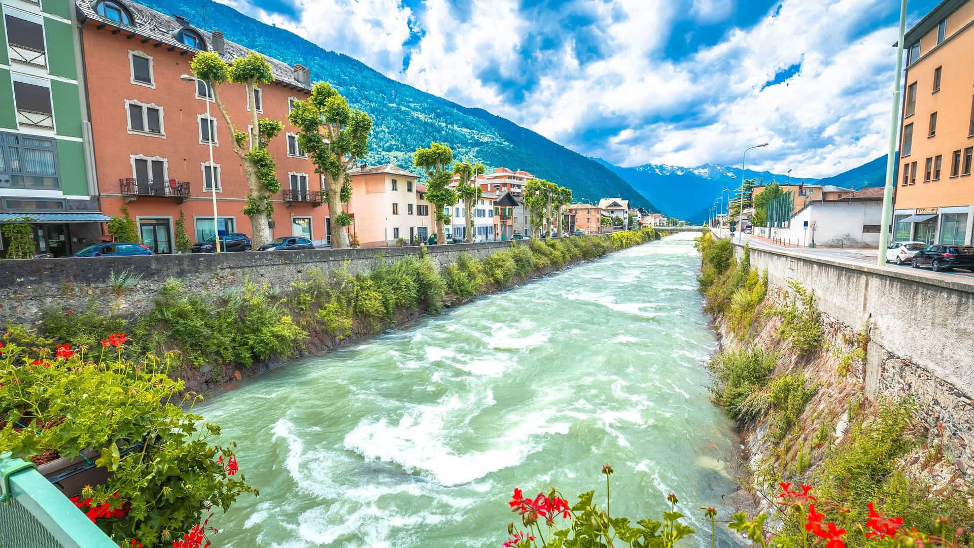 "A picturesque view of the Adda River flowing through the town of Tirano, Italy, with colorful historic buildings and the dramatic Italian Alps in the background.  "