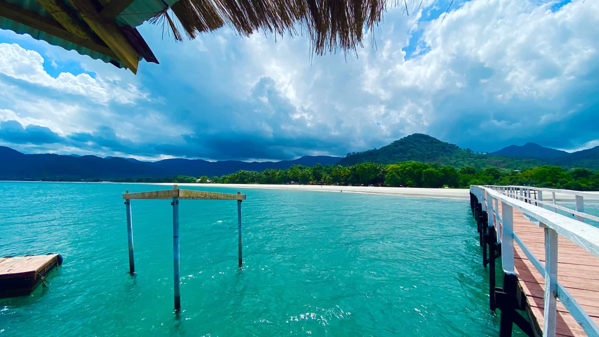 A scenic view from under a thatched roof of the stunning white-sand beach and clear turquoise water in Tokeh, Sierra Leone, with lush green hills and a white pier in the foreground.