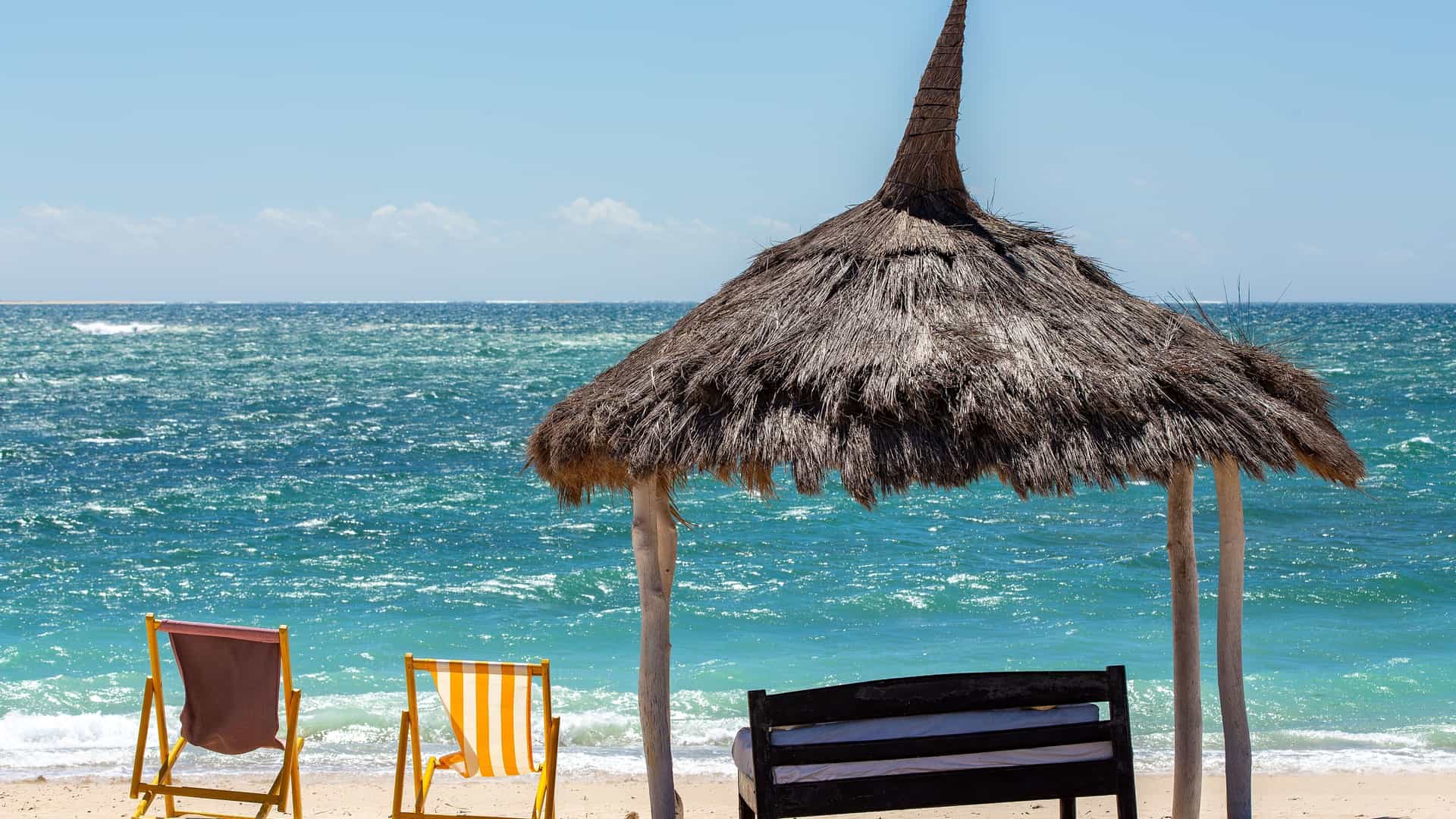 A relaxing scene on the sandy beach of Toliara (Tuléar), Madagascar, with a thatched-roof cabana and colorful lounge chairs facing the bright blue ocean.