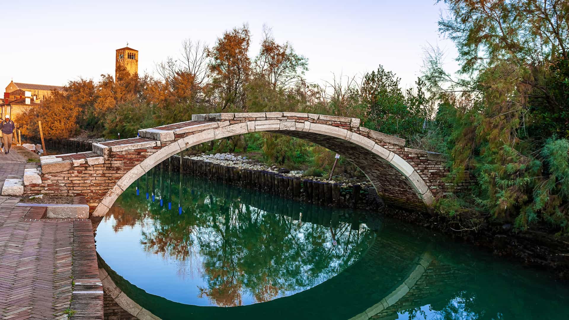 A peaceful canal scene in Torcello with a stone arch bridge forming a near‑perfect circle in its reflection, a cobblestone path where two people walk, and autumn trees and a historic bell tower glowing in warm light.