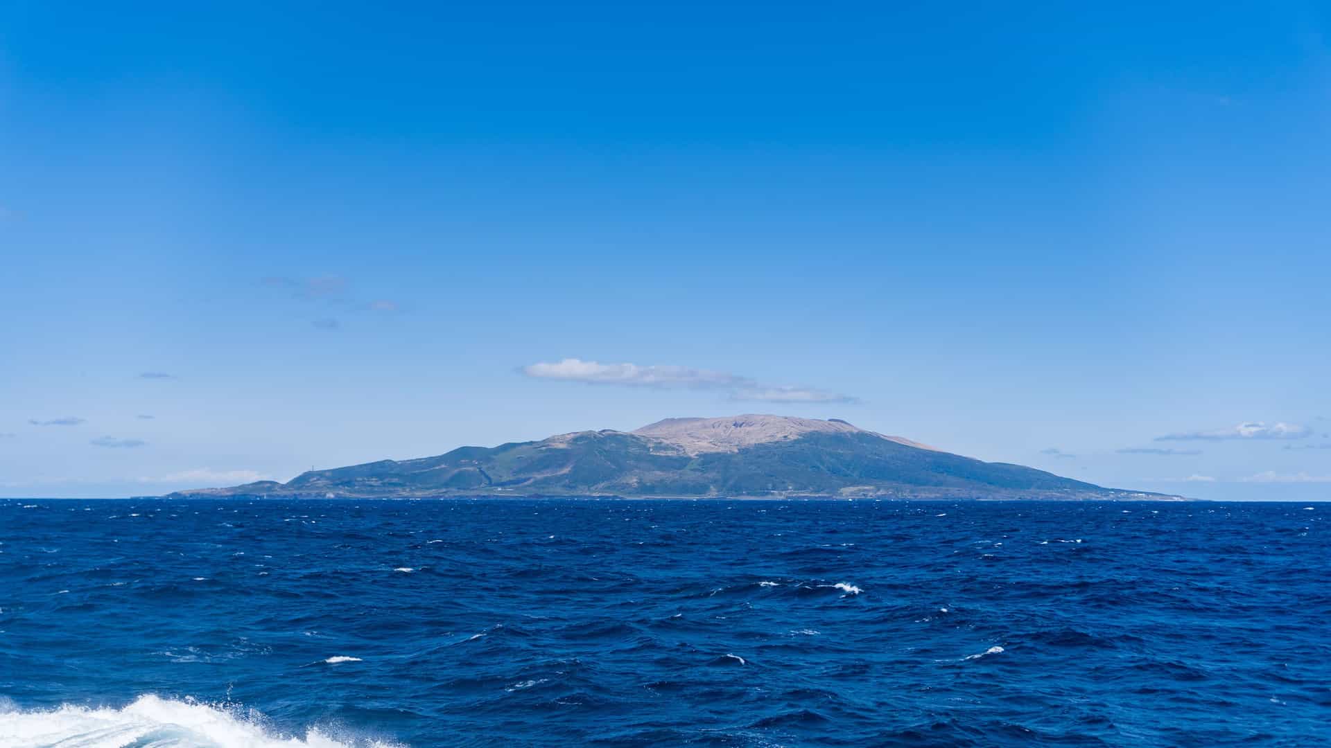 A serene view of the uninhabited volcanic island of Torishima, Japan, in the Pacific Ocean, with its dome-shaped mountain rising from the deep blue sea under a clear sky.