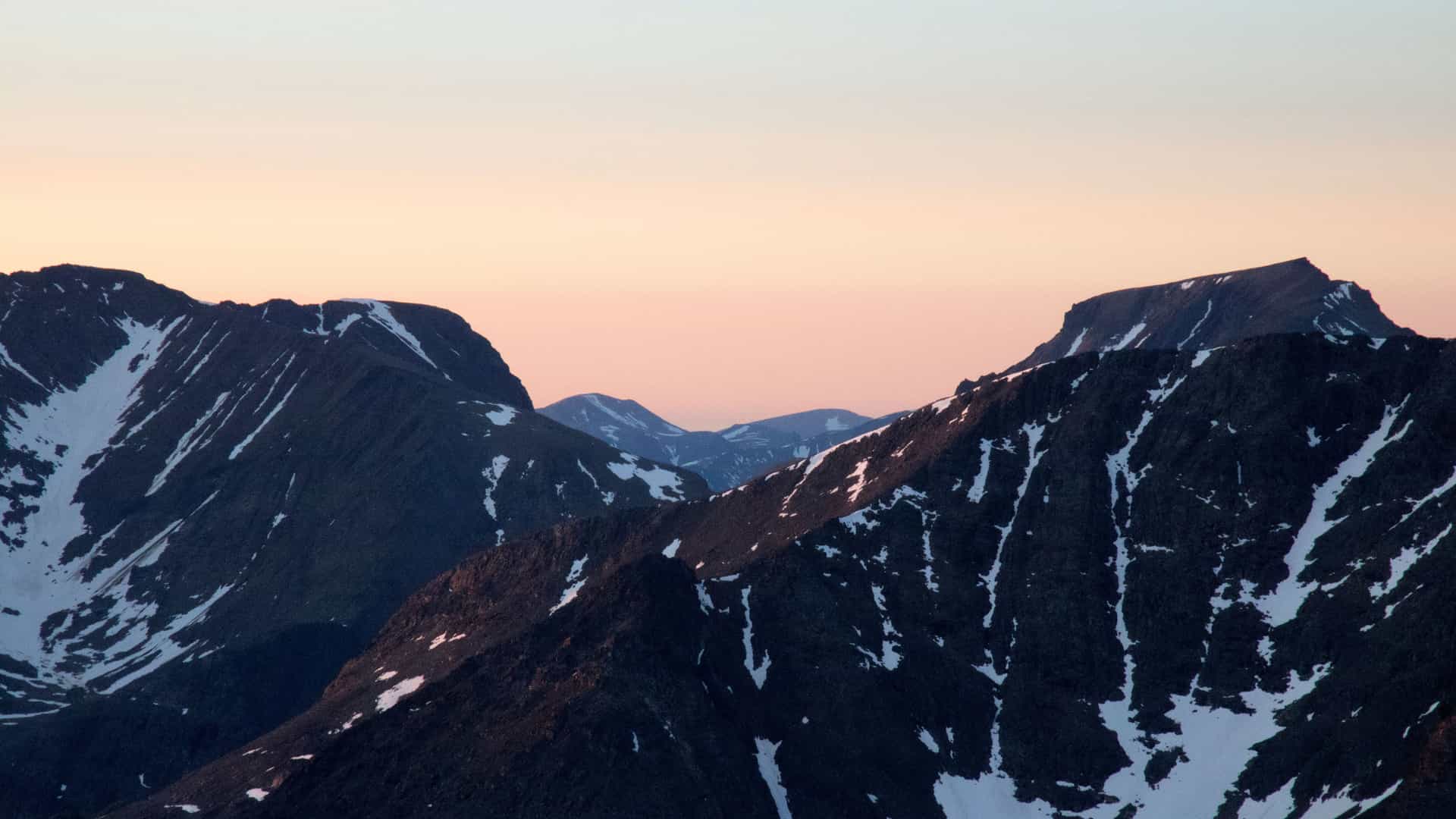 "A dramatic wide-angle shot of the rugged, snow-dusted peaks of the Torngat Mountains in Canada at twilight, with the sky fading from light pink to pale blue.  "