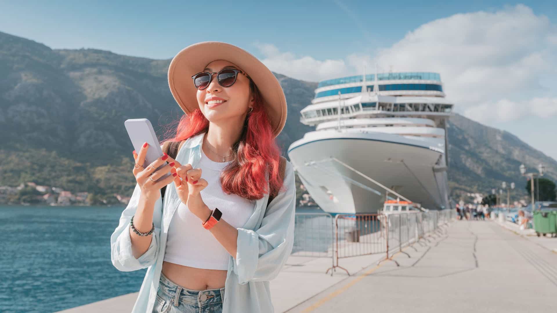 A stylish woman with a hat and sunglasses happily using her smartphone on a cruise port dock with a large white cruise ship in the background.