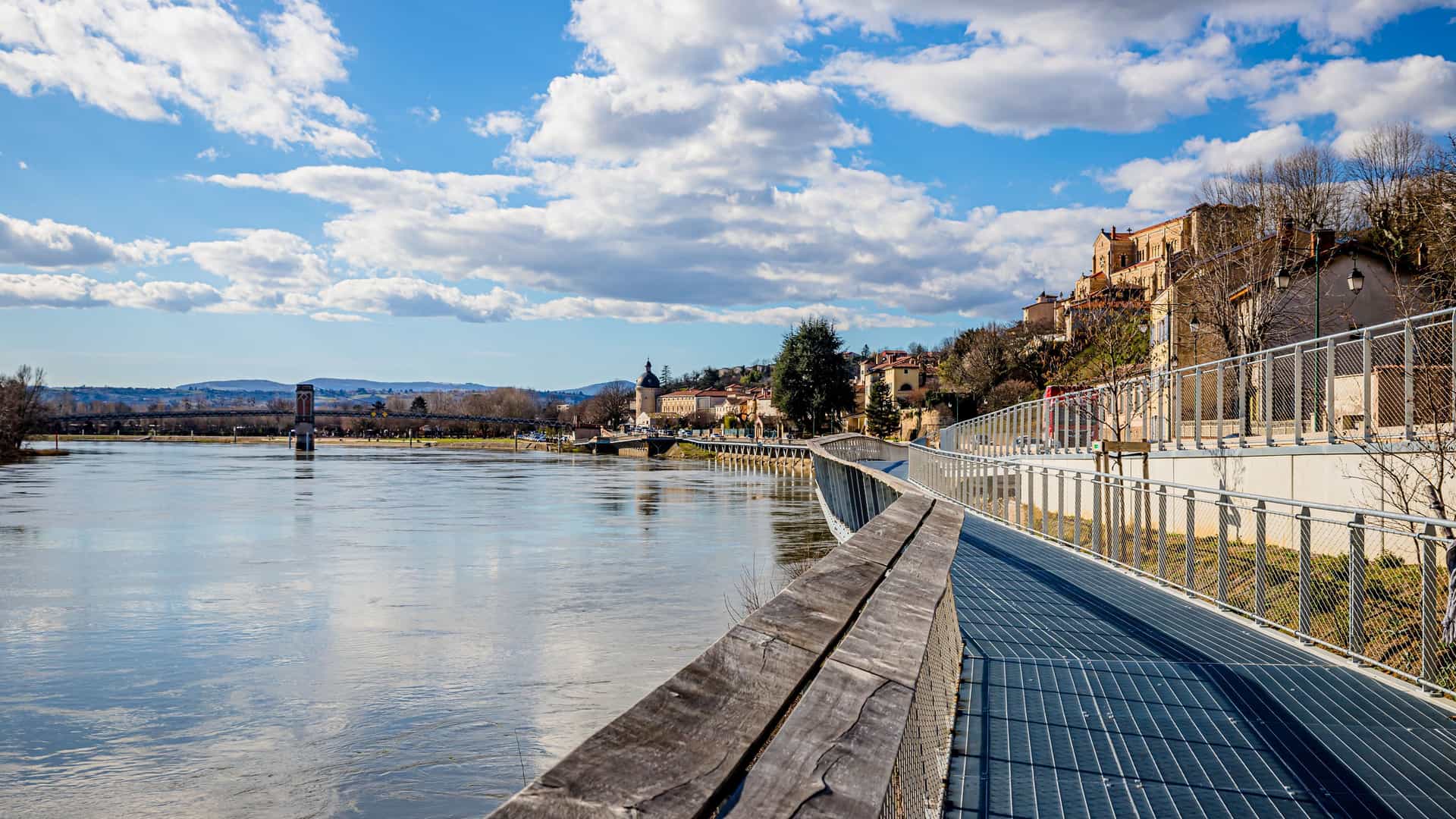 A beautiful view of the Saône River in the medieval town of Trevoux, France, with a modern, curved boardwalk and historic buildings lining the waterfront.