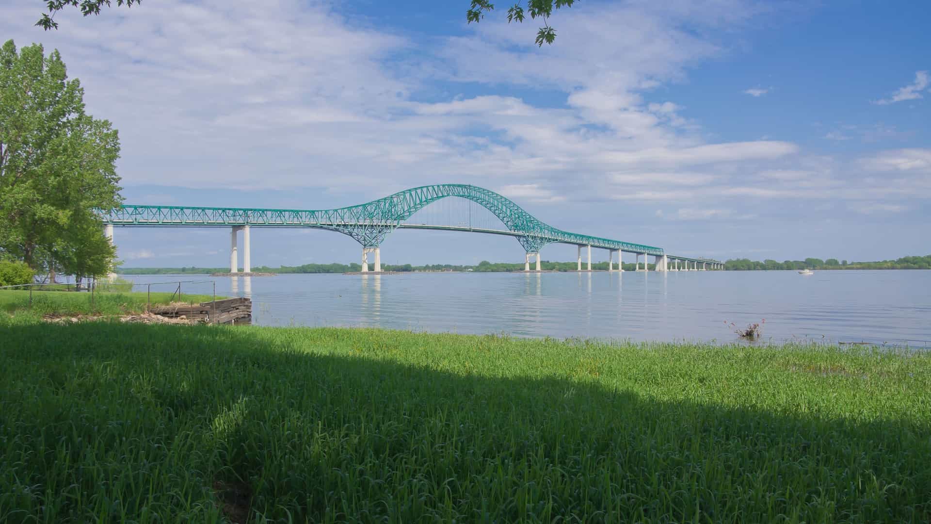 A long, green steel-arched bridge with multiple supports, the Laviolette Bridge, spanning the wide St. Lawrence River near Trois-Rivières, with a grassy riverbank in the foreground.