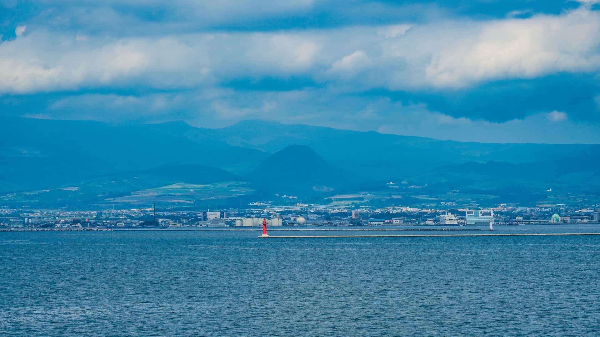 A view of the city of Hakodate, Japan, across the Tsugaru Strait, with a prominent red lighthouse on a breakwater and mountains on the horizon under a dramatic, cloudy sky.