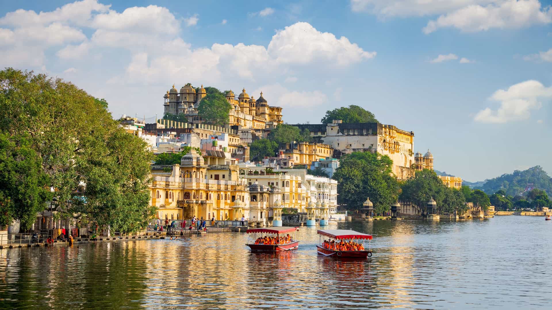 A picturesque view of the historic city of Udaipur, India, with the City Palace and other traditional buildings on the shores of Lake Pichola with two boats sailing on it.