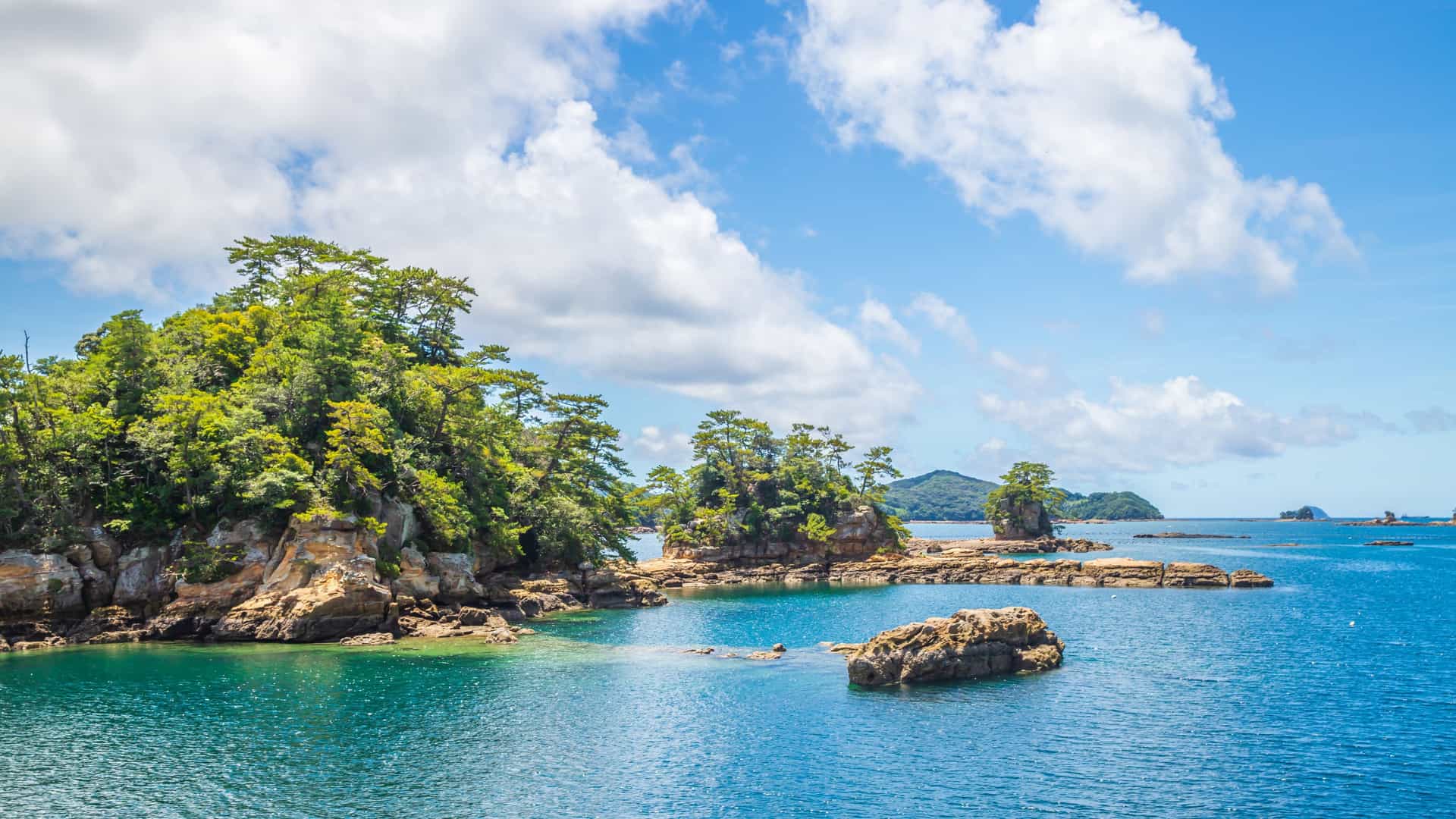 A serene coastal view in Uragashira with small rocky islets covered in lush green trees rising from vibrant blue water, all beneath a partly cloudy sky that adds softness to the tranquil scene.