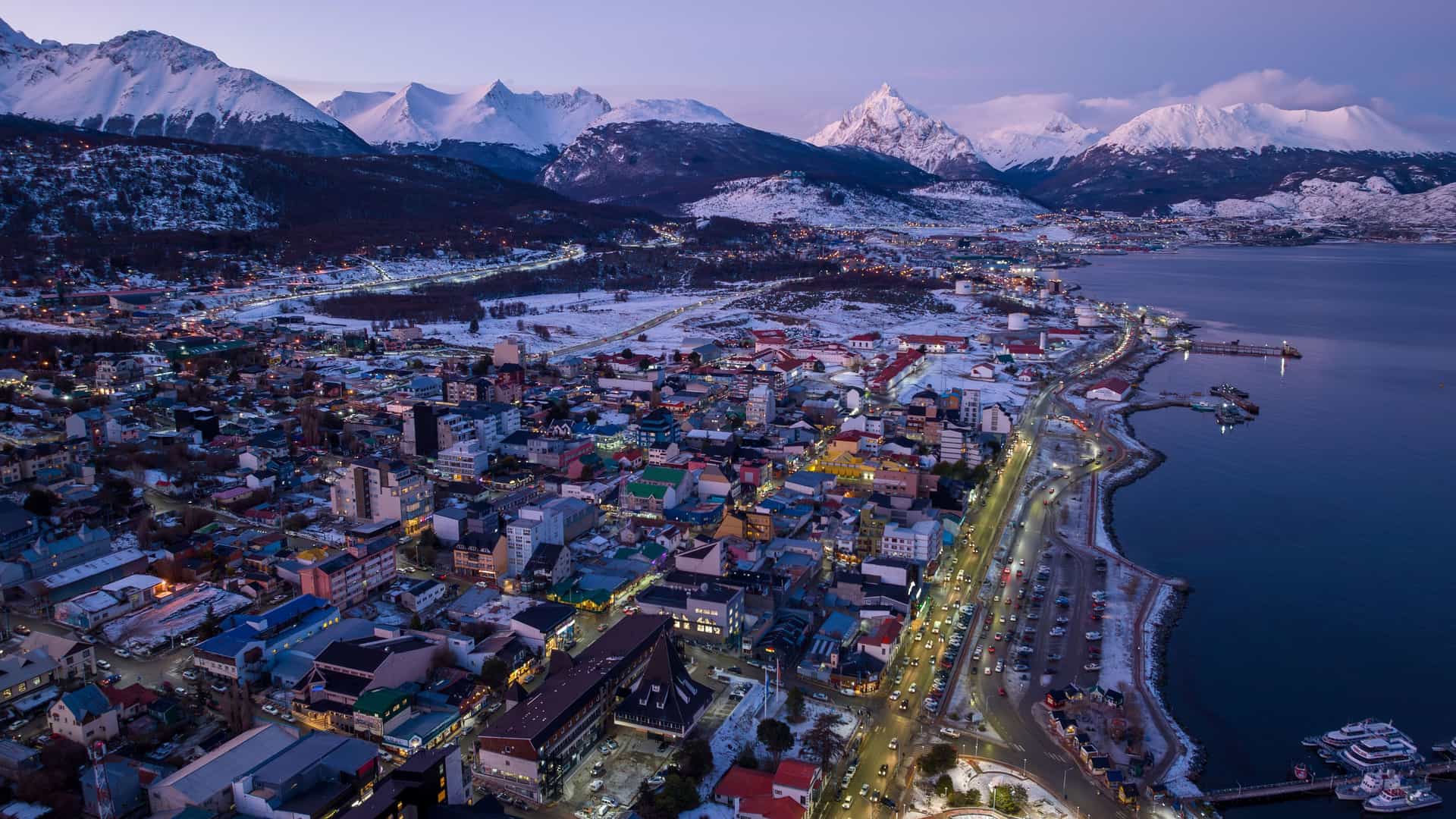 An aerial shot of the port city of Ushuaia, Argentina, at dusk, showing city lights along the Beagle Channel with the snow-capped Andes Mountains in the background. 