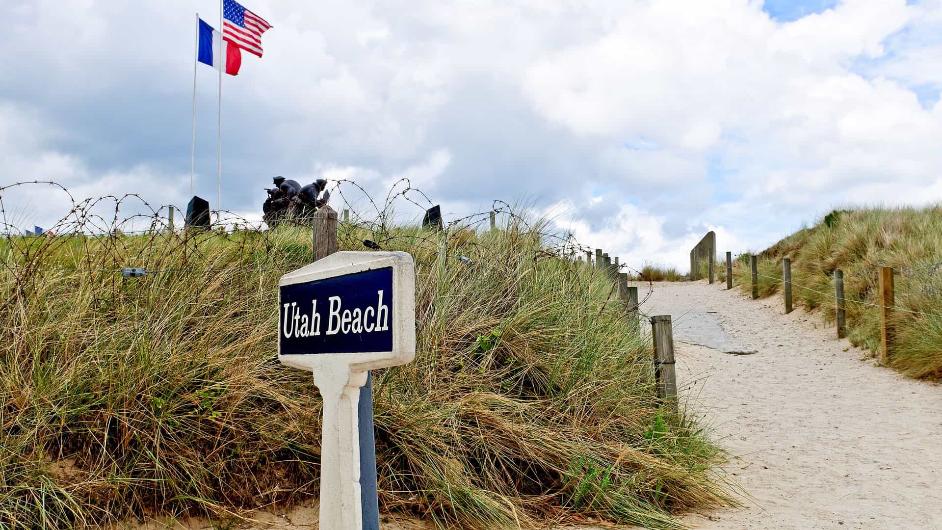 A weathered sign for 'Utah Beach' stands amidst tall grasses and barbed wire on the historic Normandy coastline, with the French and American flags waving in the distance.