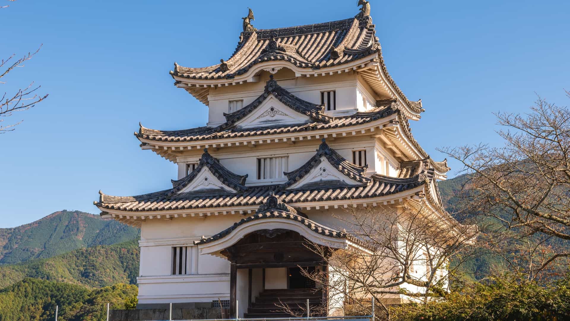 The white, multi-story Uwajima Castle keep features intricate, dark tiled roofs, set against a blue sky and forested hills.
