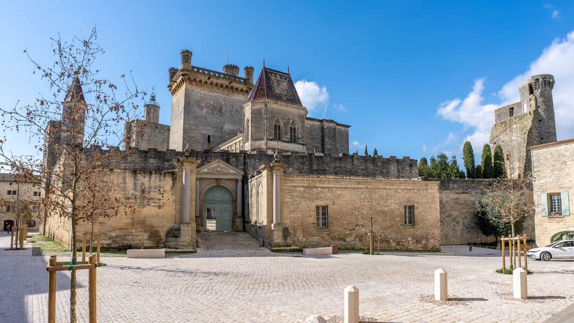 The Ducal Palace of Uzès, a medieval castle with crenelated towers, overlooks a cobblestone square under a bright blue sky.