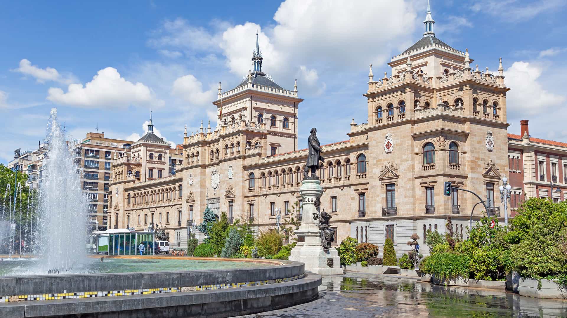 "A sunny daytime shot of the Royal Palace of Valladolid, Spain, with a large fountain in the foreground and a bronze statue in front of the ornate, historic brick building.  "