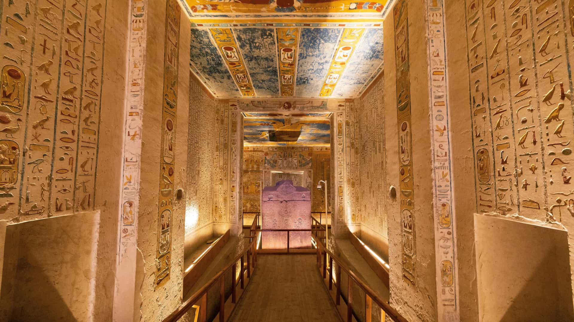 An illuminated long hallway inside an ancient Egyptian tomb in the Valley of the Kings, with walls and ceilings covered in hieroglyphs and colorful, intricate paintings.