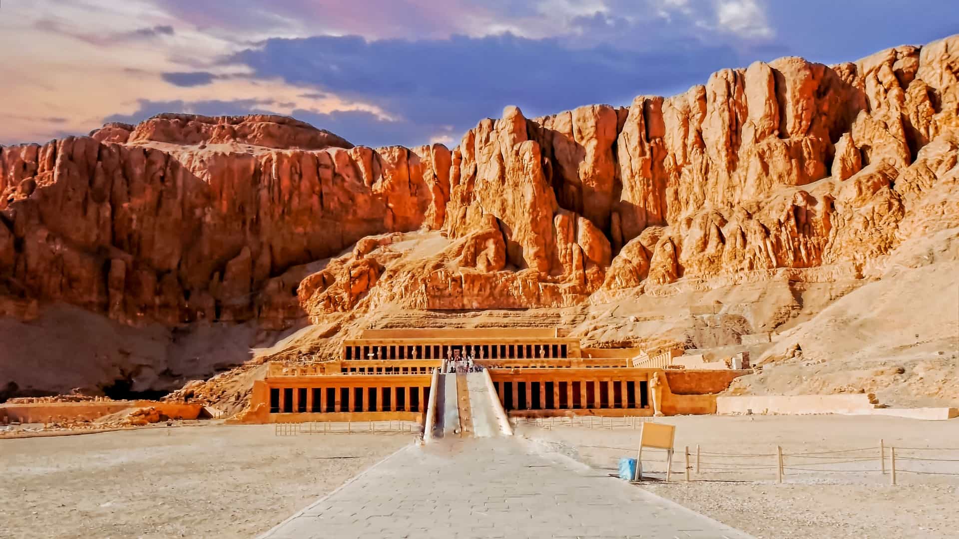 The Temple of Hatshepsut, a tiered structure, is built into the base of a towering, sheer orange cliff face under a dramatic sky.