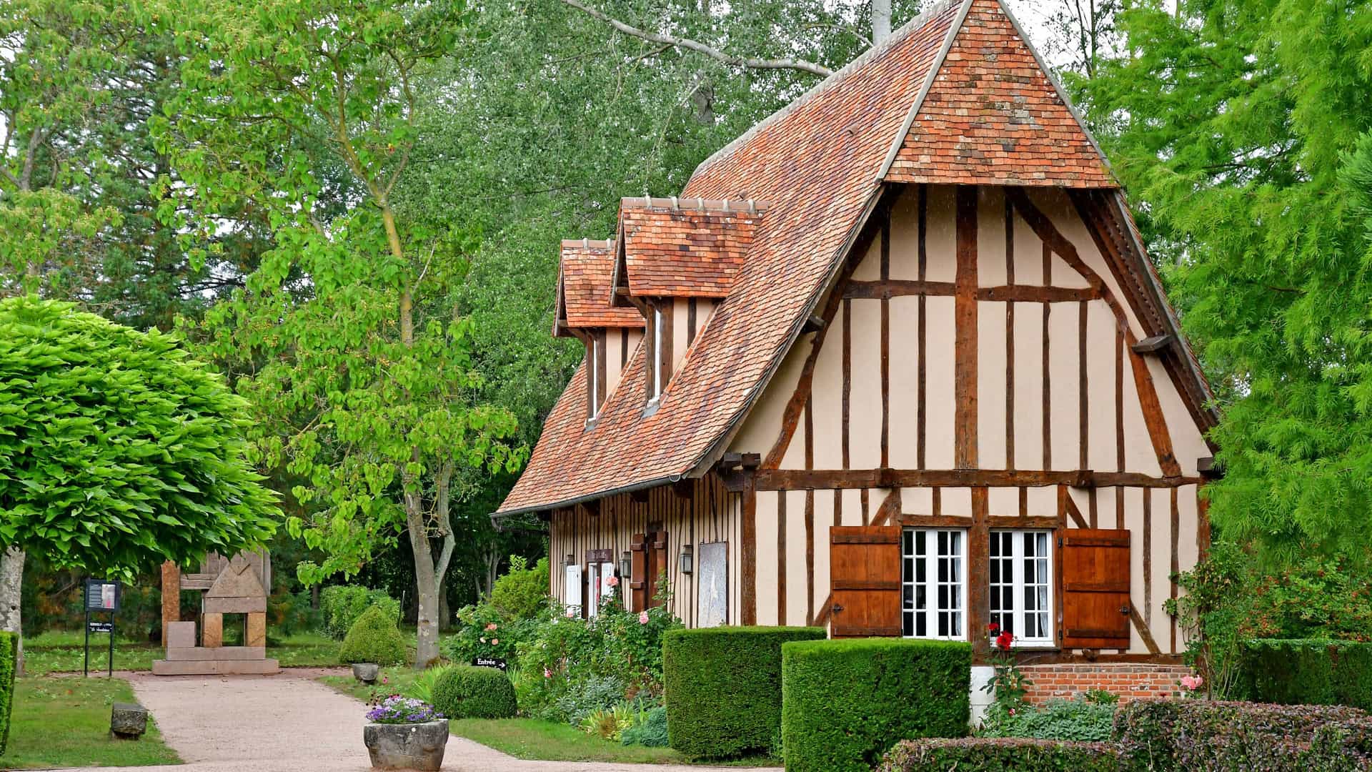 A picturesque half-timbered house with a red tiled roof and brown shutters, surrounded by lush green gardens and trees, at the Château de Vascoeuil in France.
