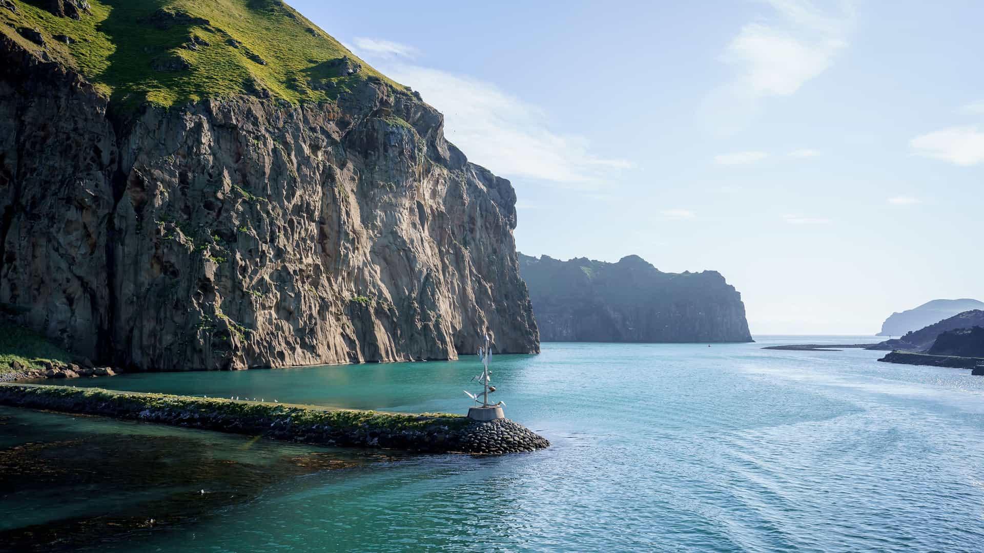 A dramatic landscape view of the harbor entrance at Vestmannaeyjar in Iceland, with steep, green-covered cliffs towering over the turquoise blue water of the bay.