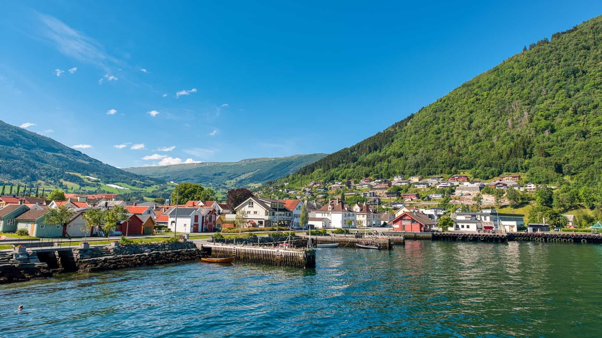 A scenic view of the village of Vik, Norway, situated in a green valley on the southern shore of the Sognefjord, with colorful buildings clustered near the water and surrounded by steep, rugged mountains.