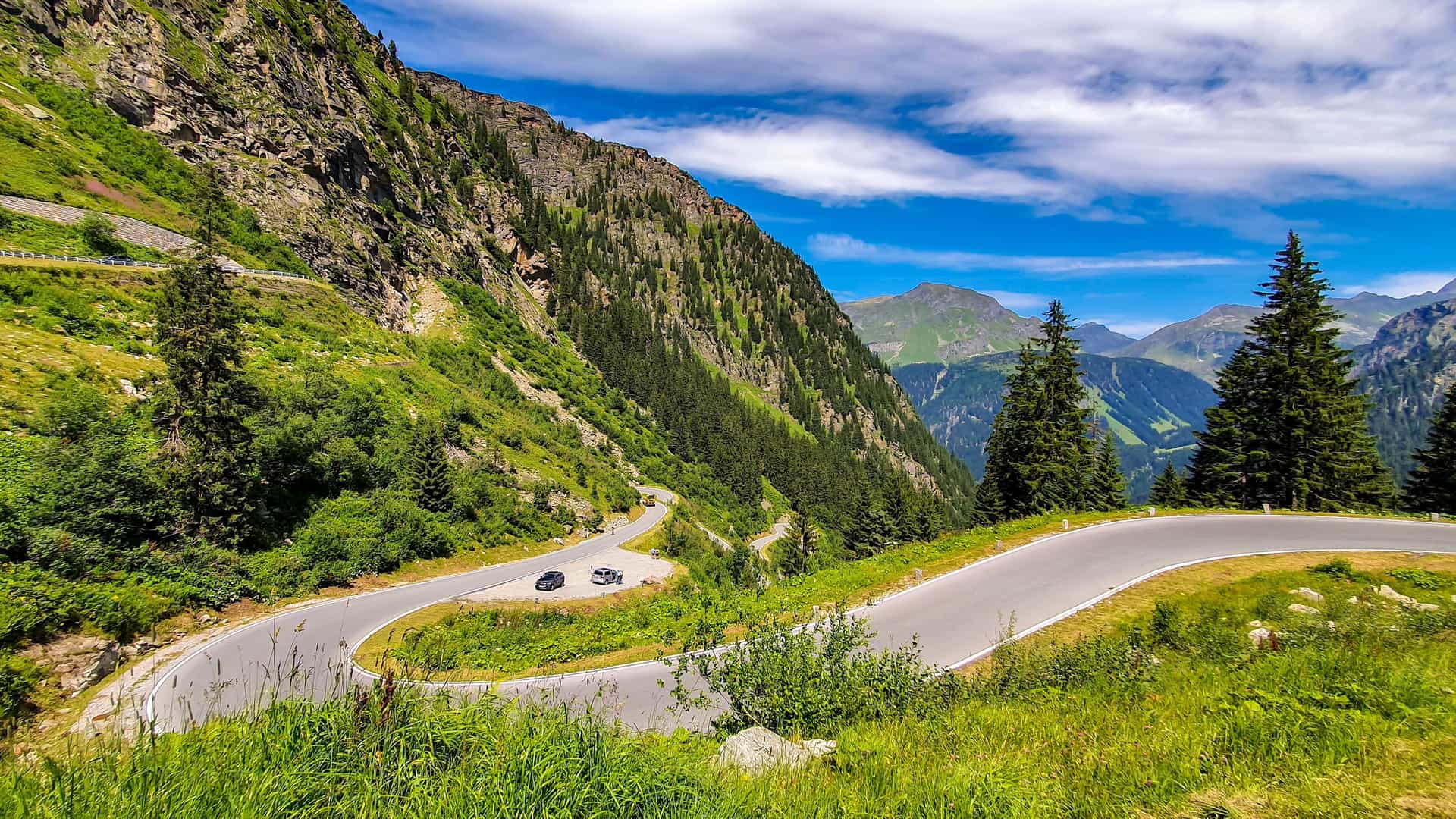 A stunning aerial shot of the Silvretta High Alpine Road, a winding, scenic mountain pass in Vorarlberg, Austria, with cars on the road and dramatic green mountains in the background.