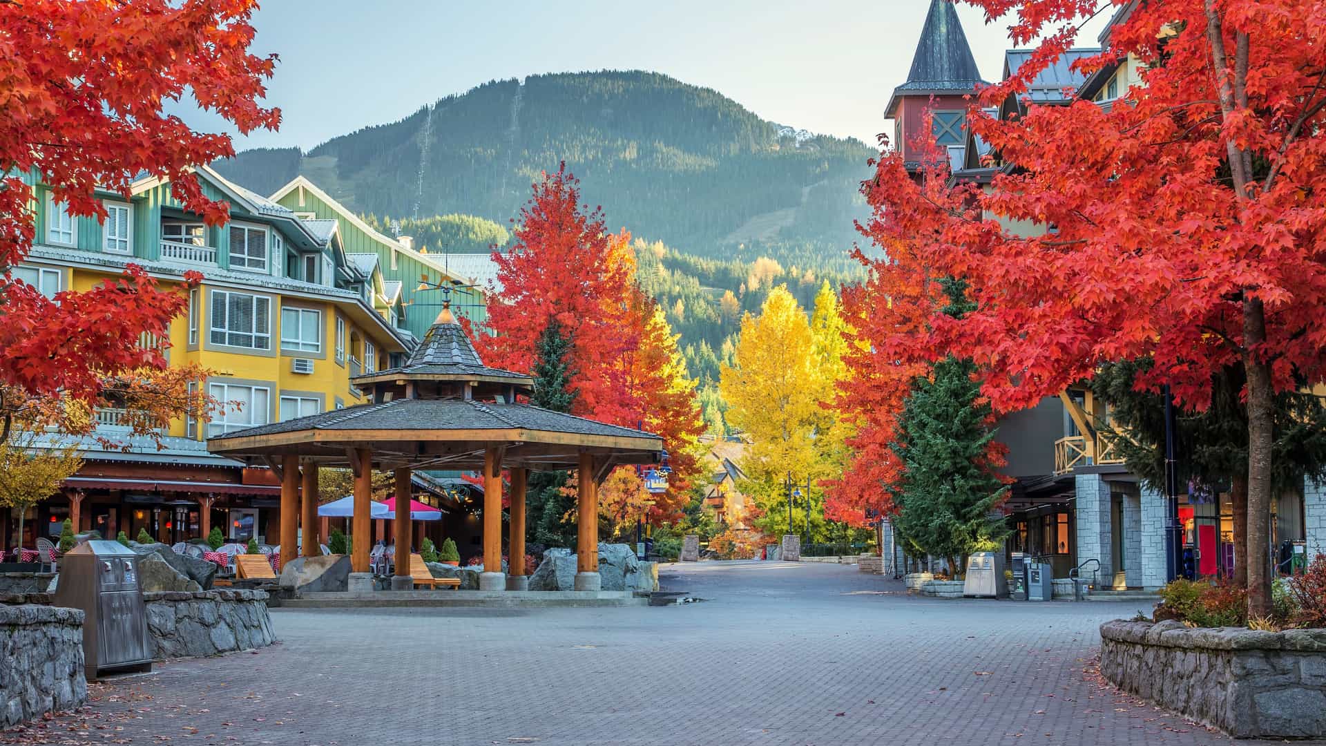 "A scenic view of Whistler Village Square in autumn, with colorful buildings, a central gazebo, and trees with vibrant red and yellow foliage against a backdrop of mountains.  "