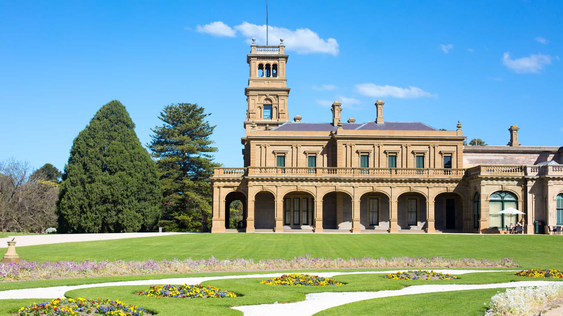 A stunning view of the historic Victorian-era Werribee Mansion in Wyndham, Australia, with its grand architecture, manicured gardens, and a bright blue sky with white clouds.