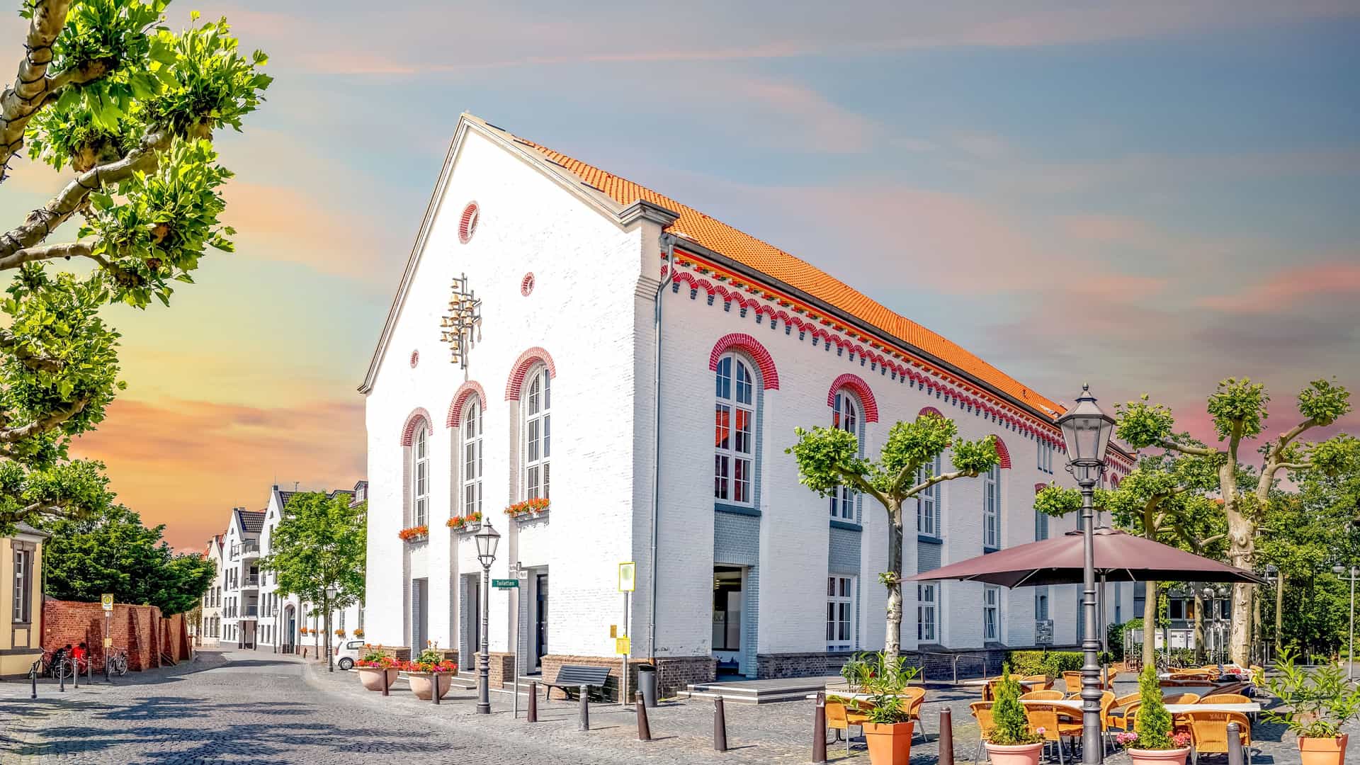 A wide, cobbled street with a large, white building with a red tile roof and arched windows. Tables and chairs are outside under an umbrella, and mature trees line the street in Xanten, Germany.