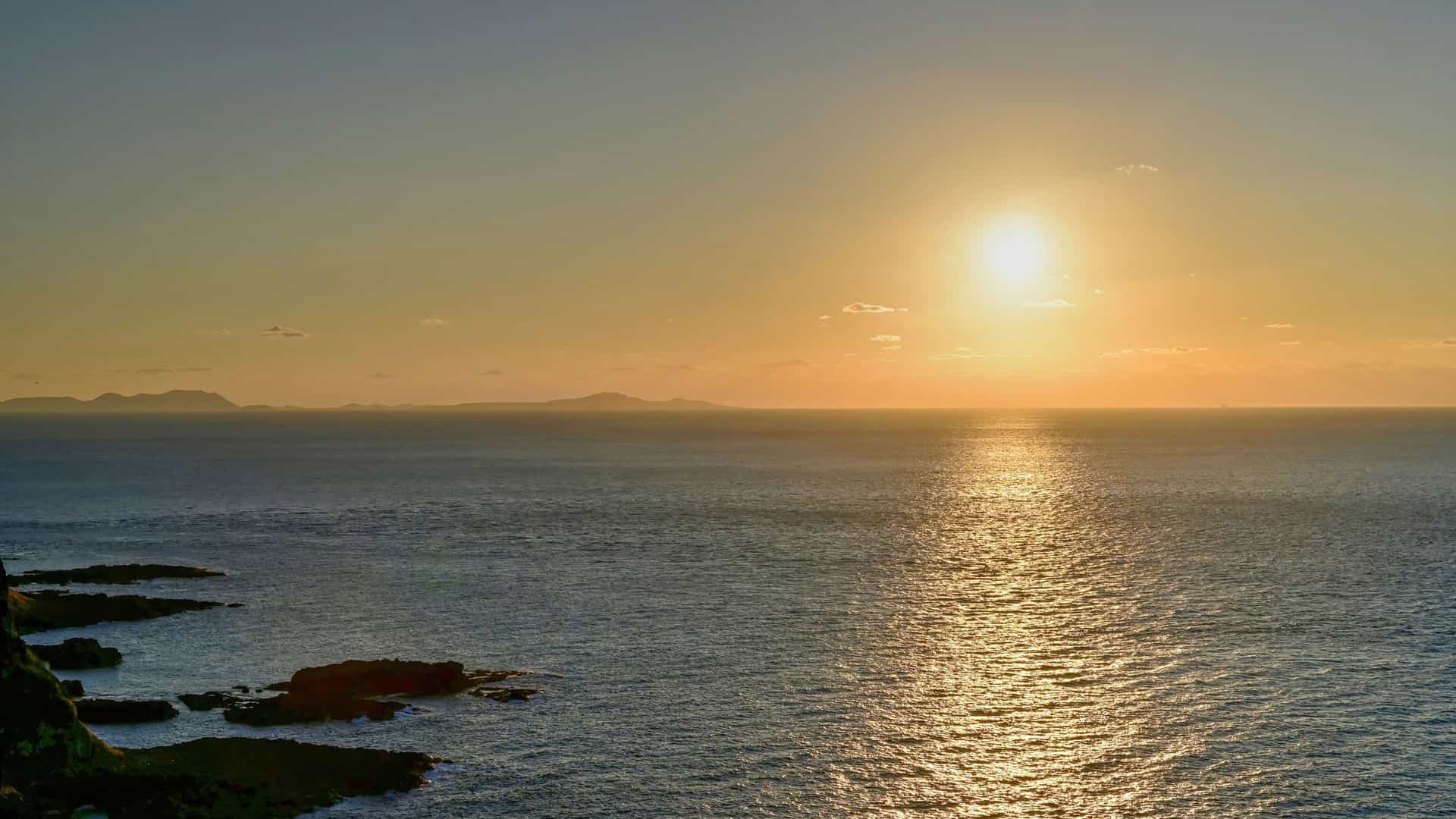 A golden sunset reflecting on the calm waters where the Yellow Sea meets the East China Sea, with a rocky coastline on the left and distant islands on the horizon.