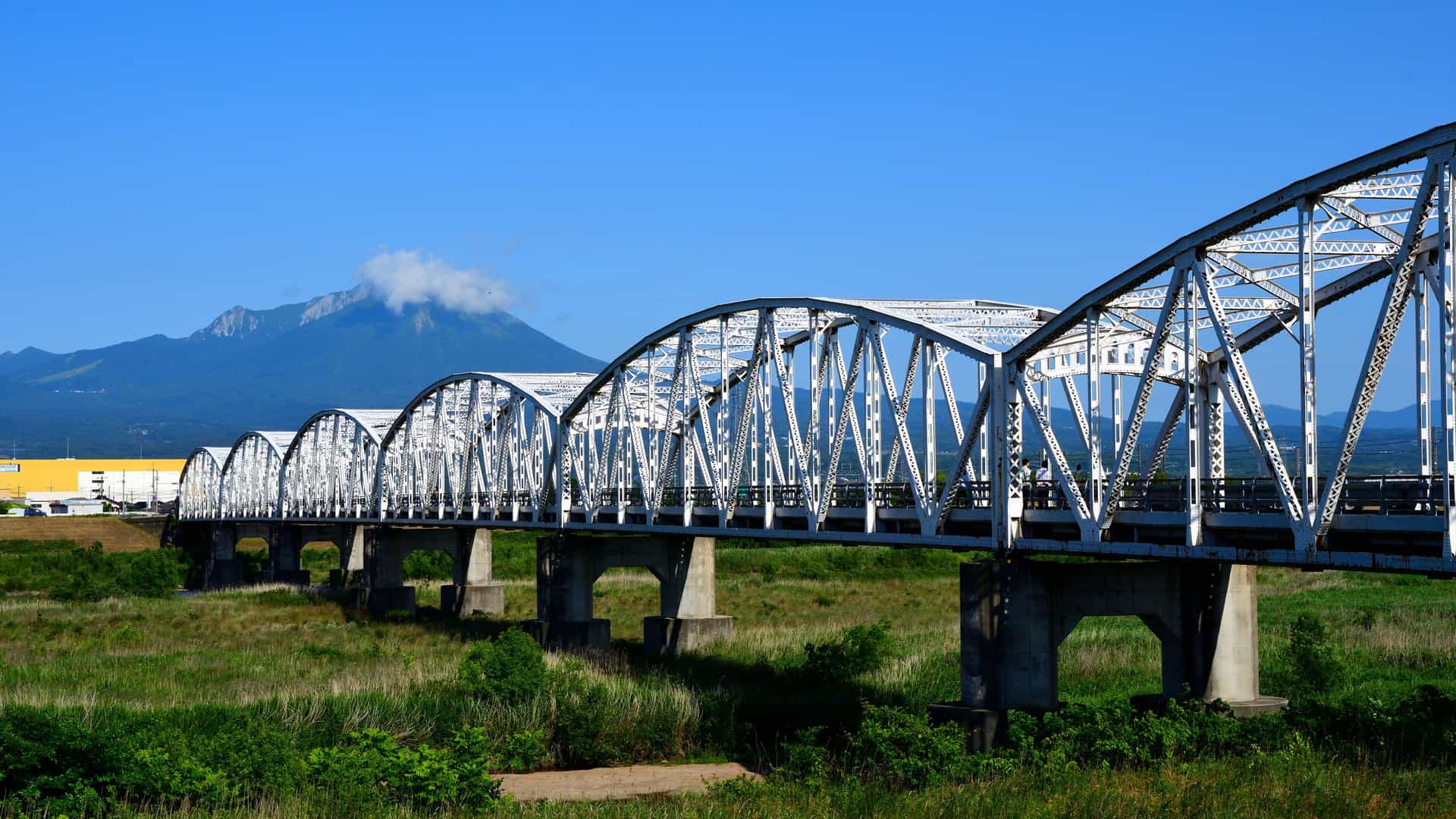 "A large white truss bridge spans a grassy field, with a prominent volcanic mountain in the background under a clear blue sky, near the city of Yonago, Japan."