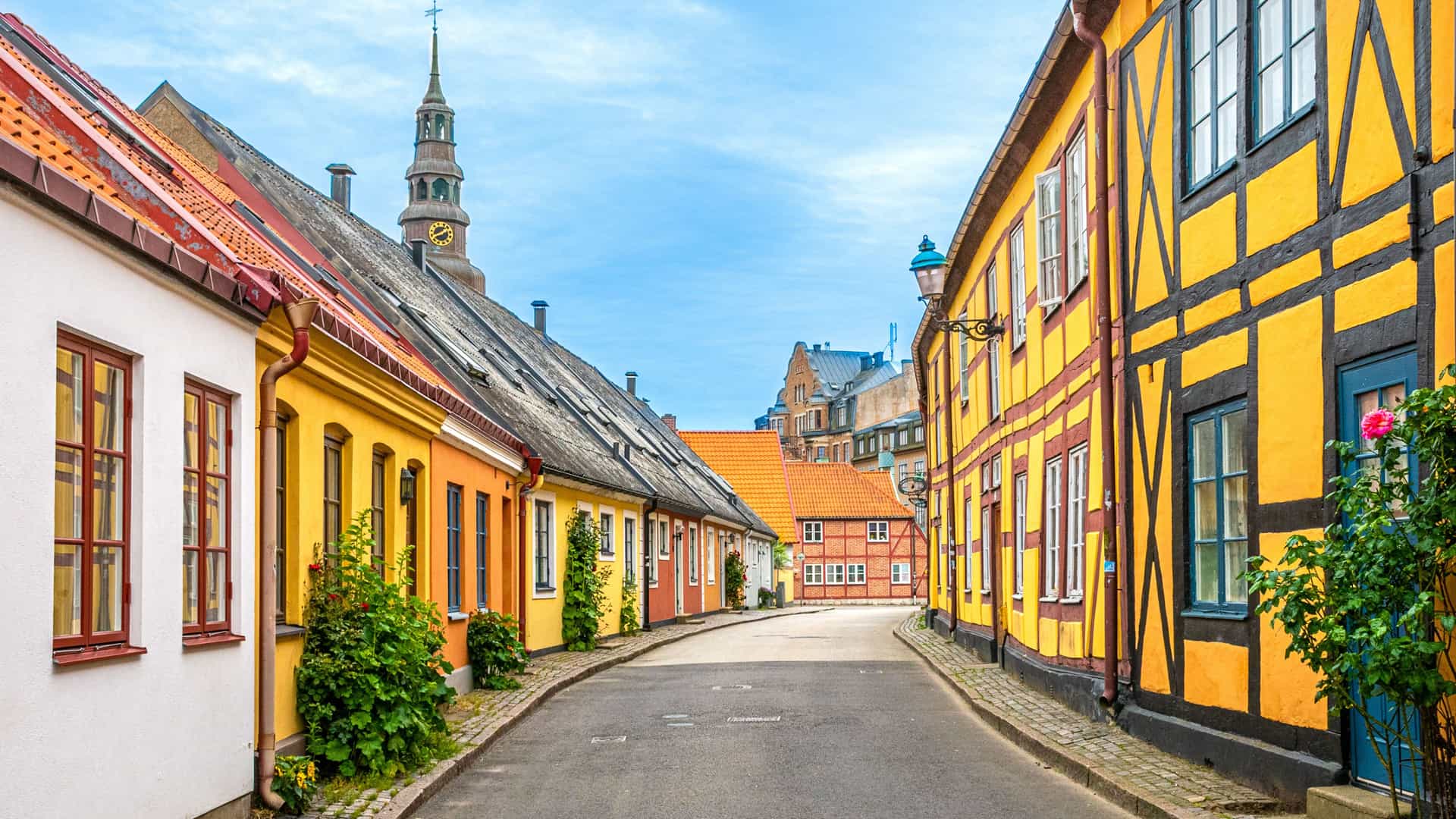 A cobblestone street curves between colorful, half-timbered buildings in the historic town of Ystad, Sweden, with a church steeple visible in the background.