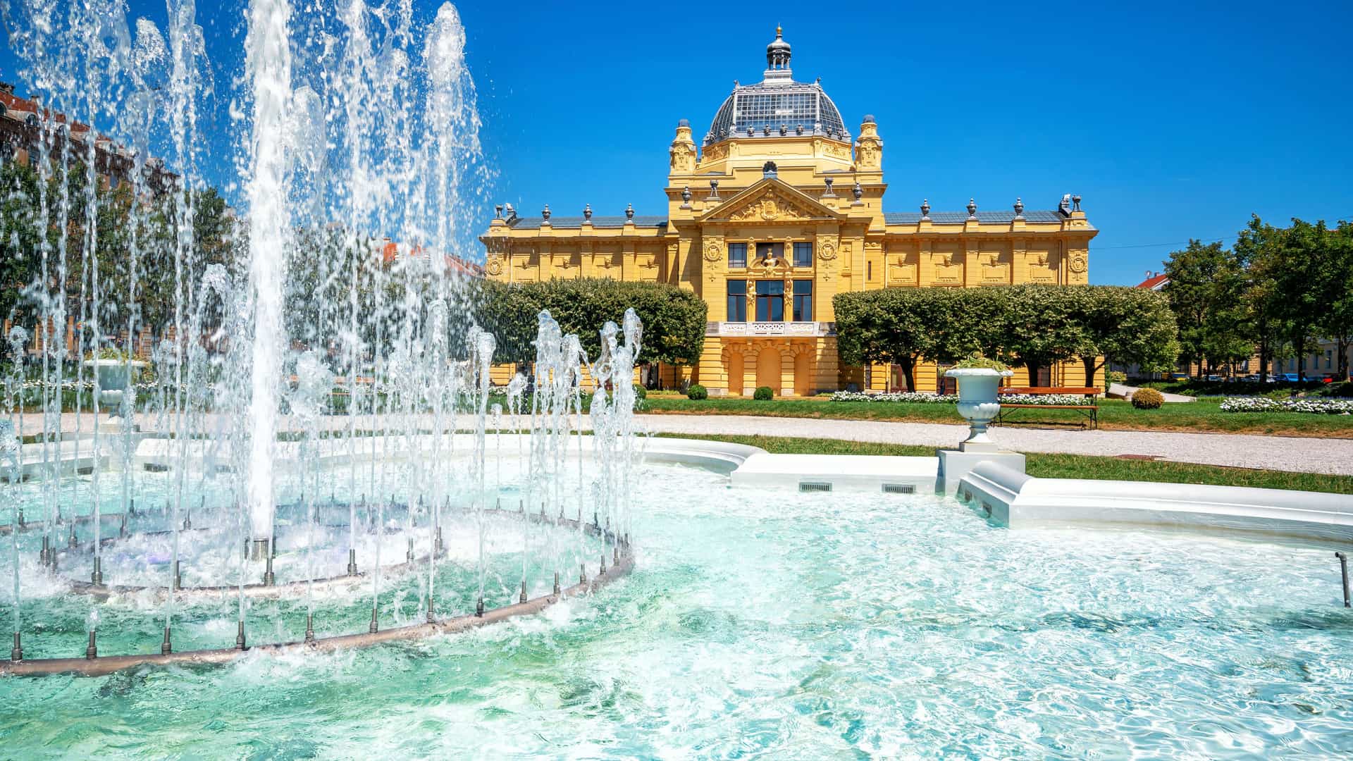 "A bright, sunlit view of the Croatian Museum of Arts and Crafts in Zagreb, with a large, bubbling fountain in the foreground and a clear blue sky overhead."