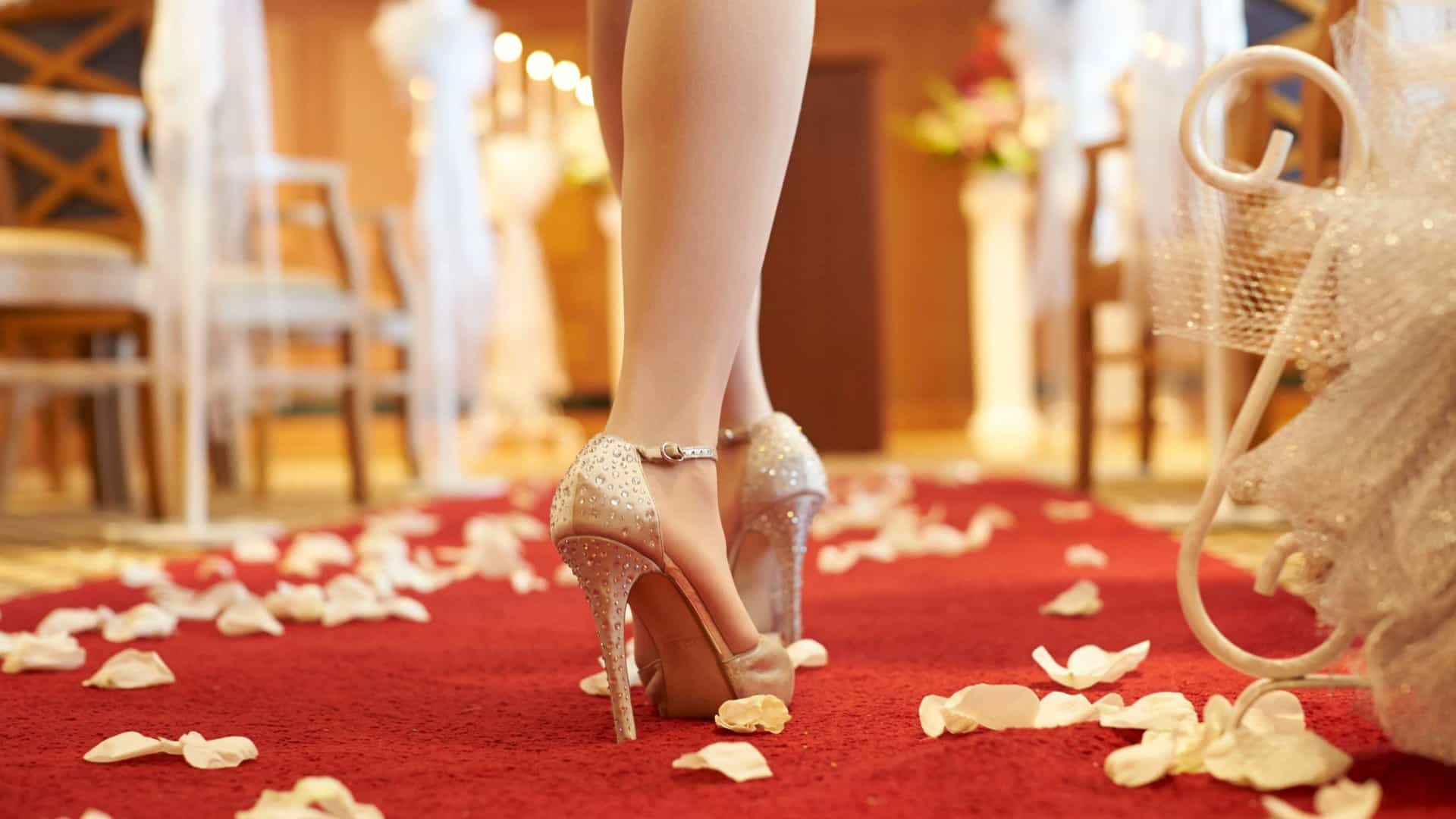 A close-up view from behind of a bride's legs and sparkling, rhinestone-embellished high heels as she stands on a red aisle runner scattered with white flower petals in a wedding chapel.