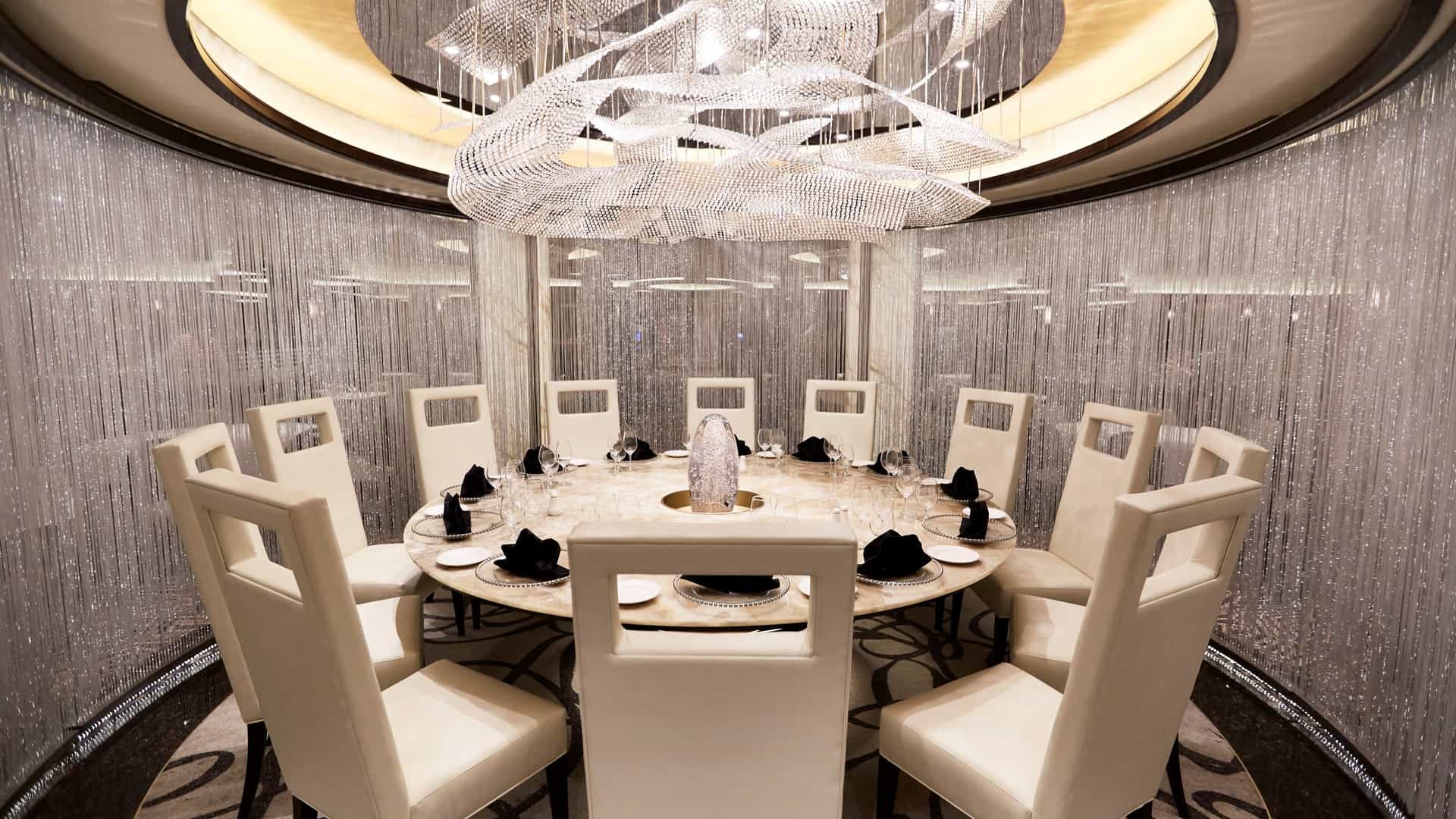 A luxurious, custom-crafted round dining table enclosed by shimmering silver beaded curtains beneath a large, elaborate crystal chandelier for an intimate private dining experience.