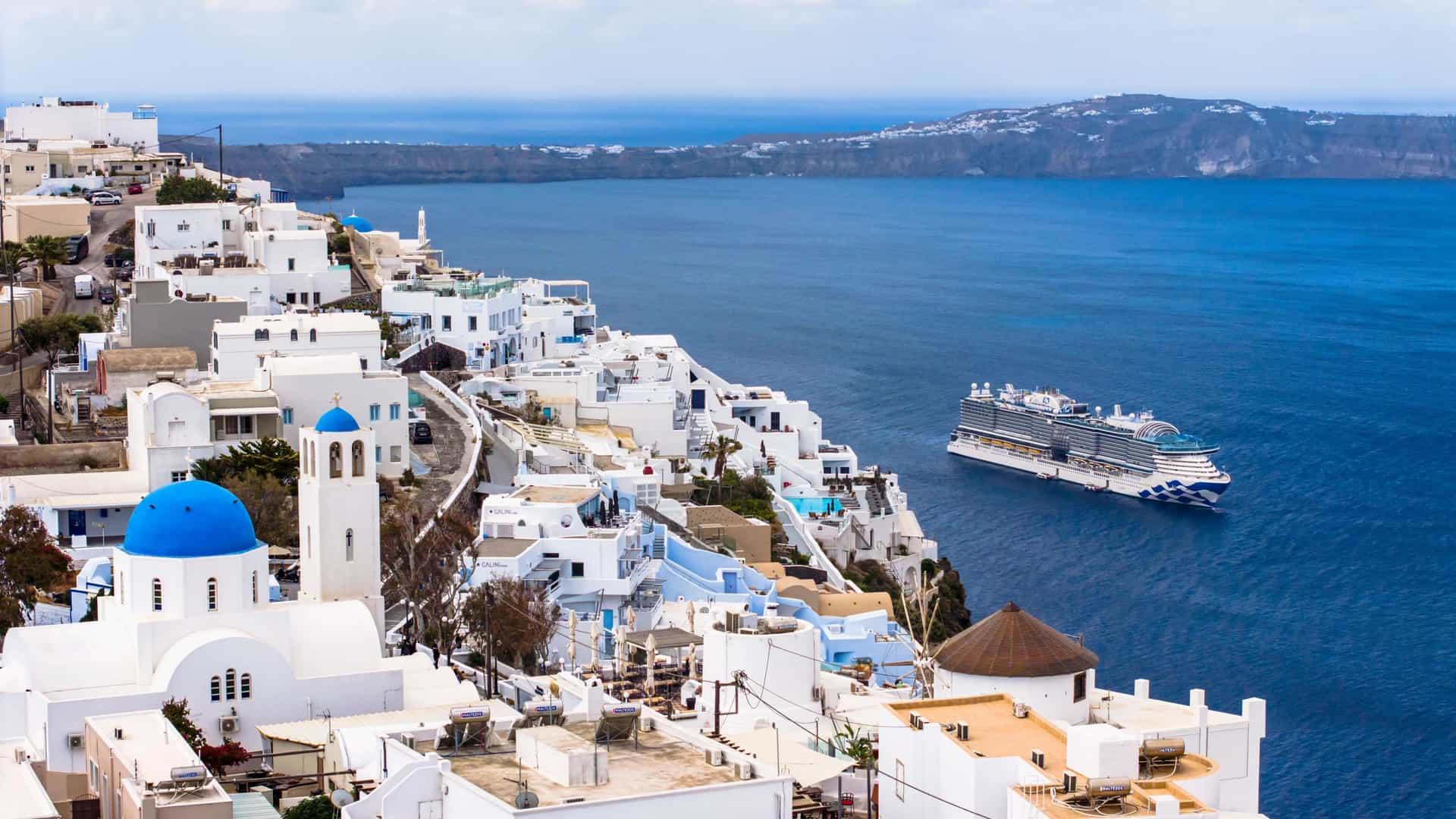 An iconic aerial view of Santorini, Greece, featuring white-washed buildings, blue domes, and a Princess Cruises ship anchored in the deep blue Mediterranean caldera.