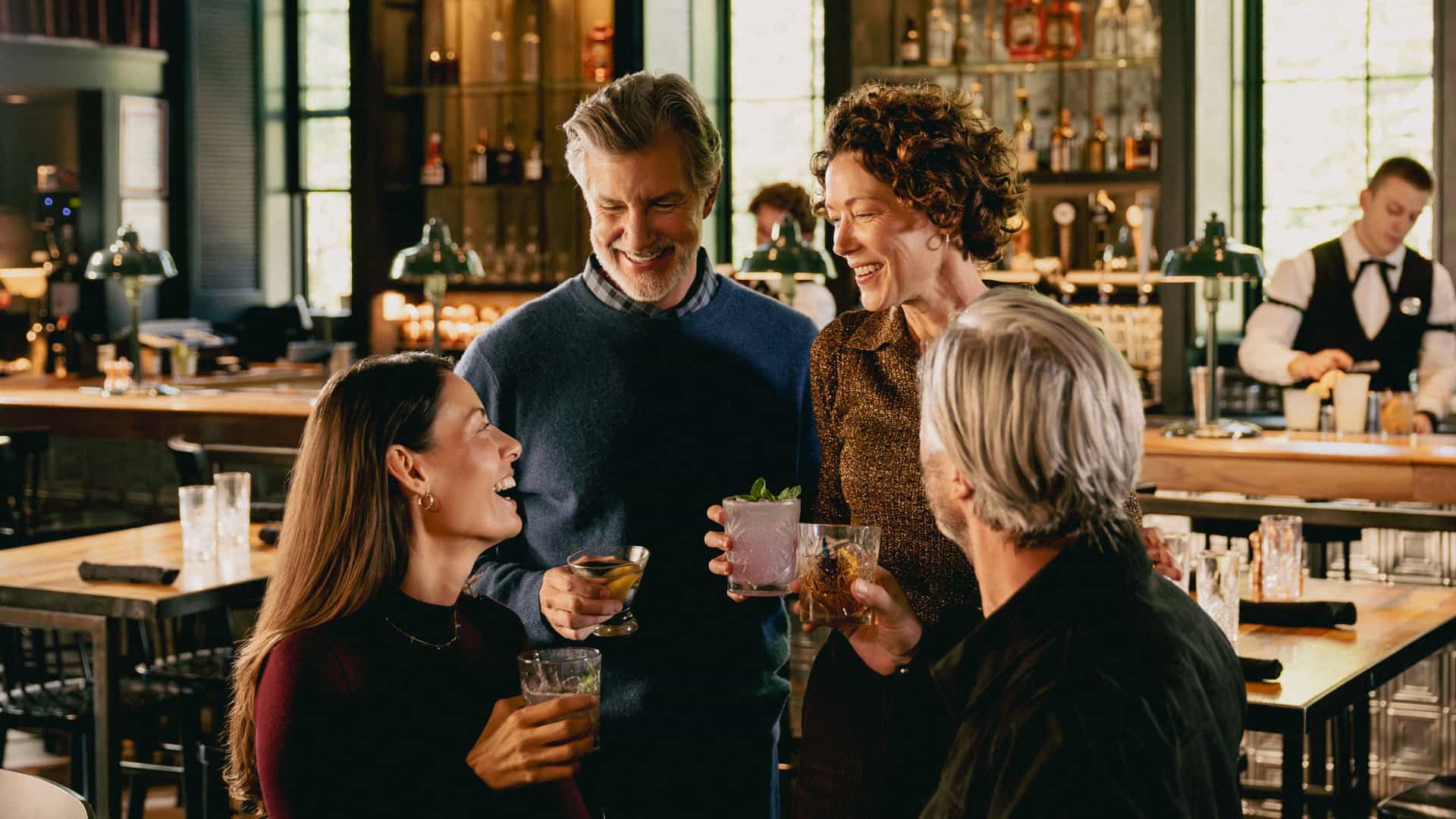 Four adults standing at a warmly lit bar, smiling and chatting while holding cocktails, with a bartender and shelves of bottles in the background.